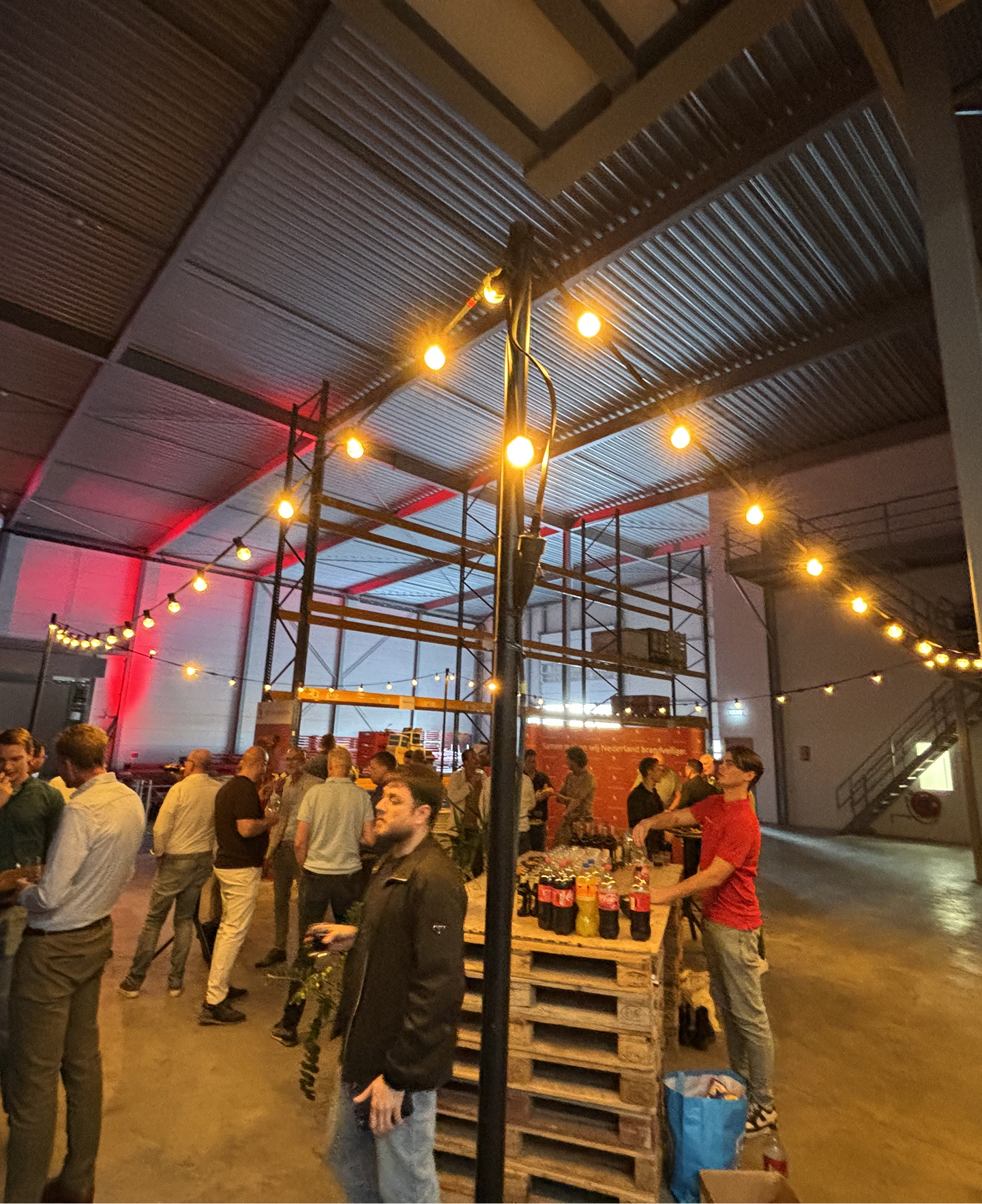 Group of people socializing at an indoor event with string lights and a table made of stacked wooden pallets holding drinks.