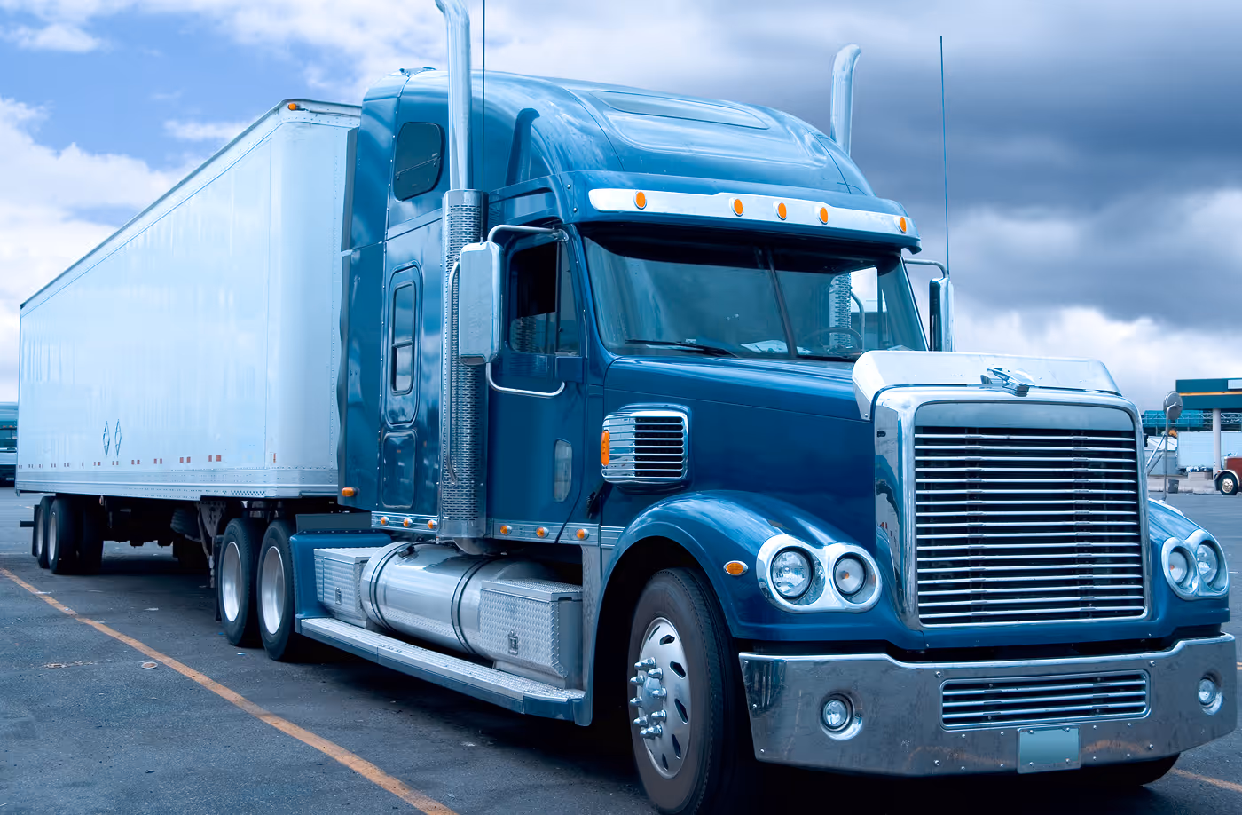 Blue semi-truck with white trailer parked on asphalt under a cloudy sky.