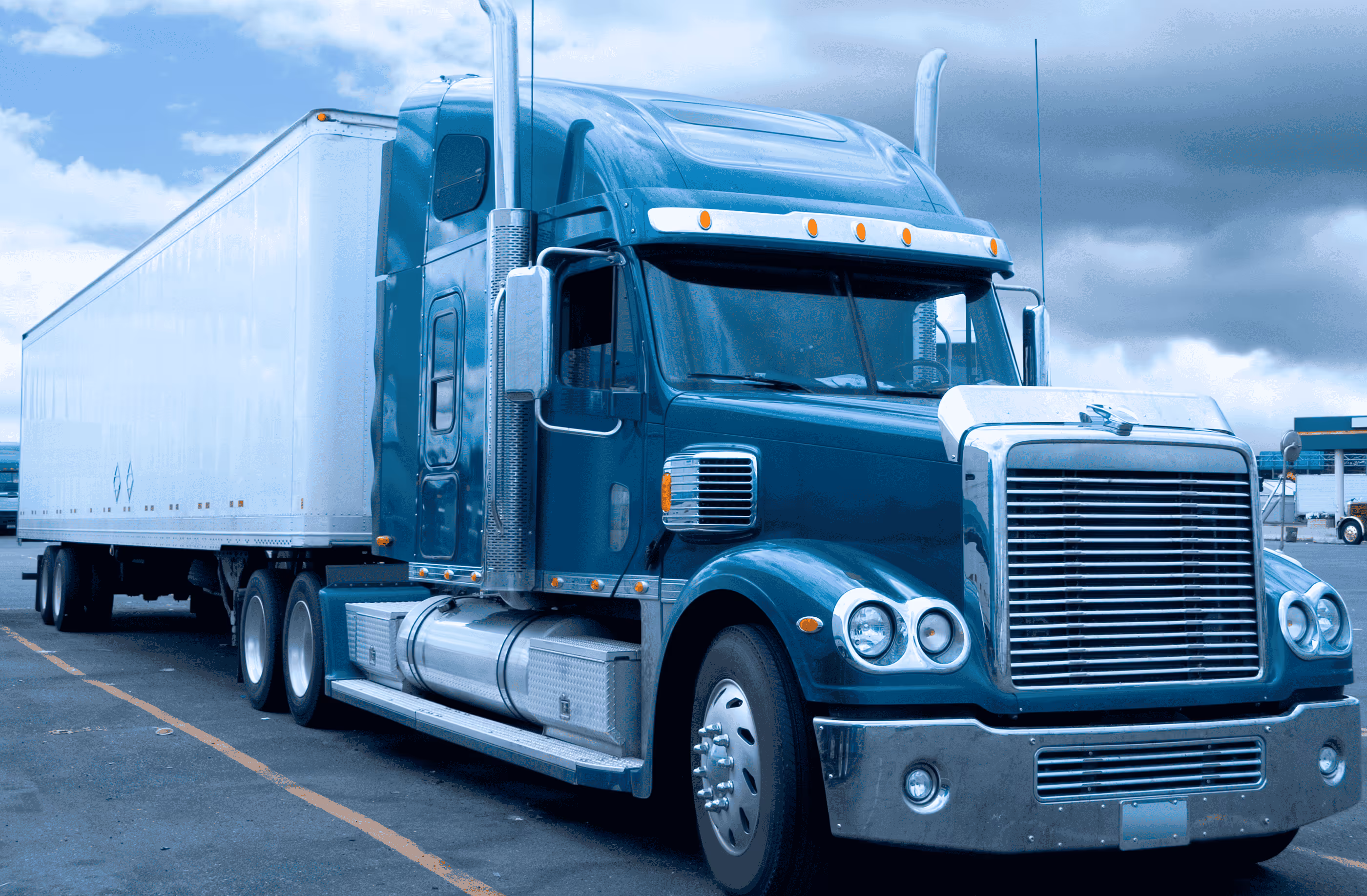 Blue semi-truck with white trailer parked on asphalt under a cloudy sky.