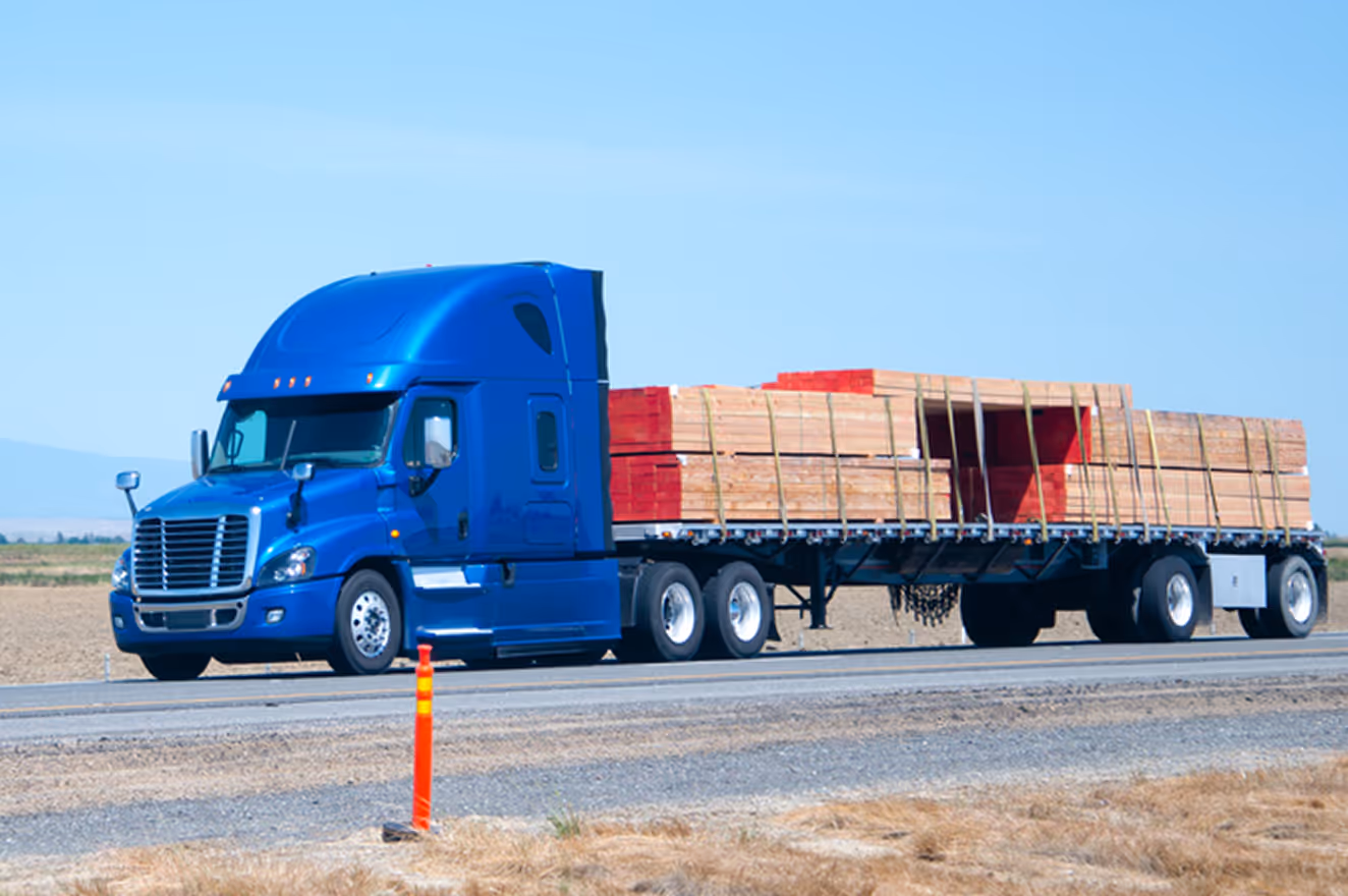Blue semi-truck hauling flatbed trailer loaded with strapped wooden planks on highway.