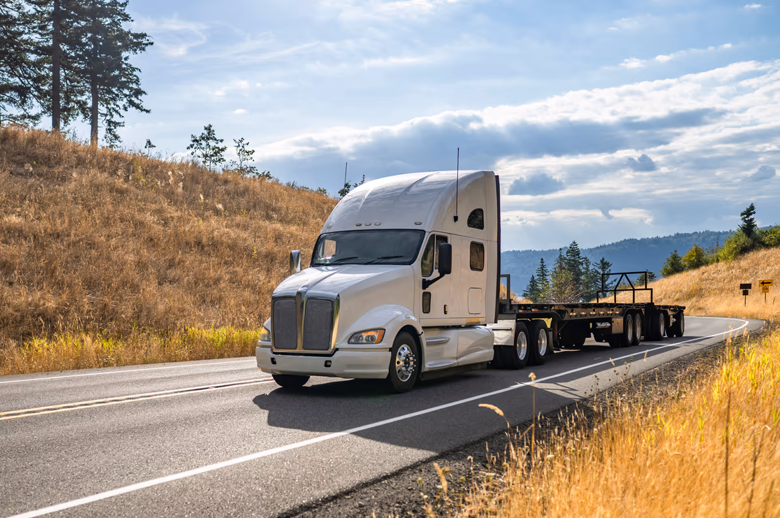 White semi truck with empty flatbed trailer driving on a rural road by dry grass and hills.