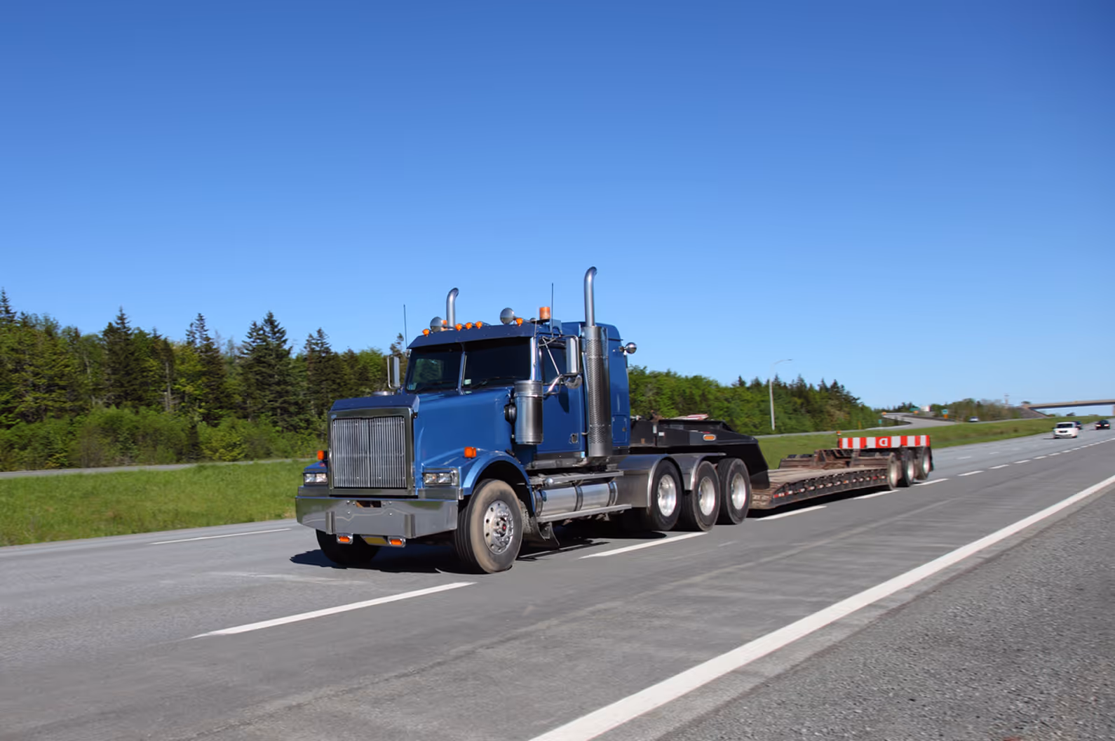 Blue semi-truck with empty flatbed trailer driving on highway under clear blue sky.