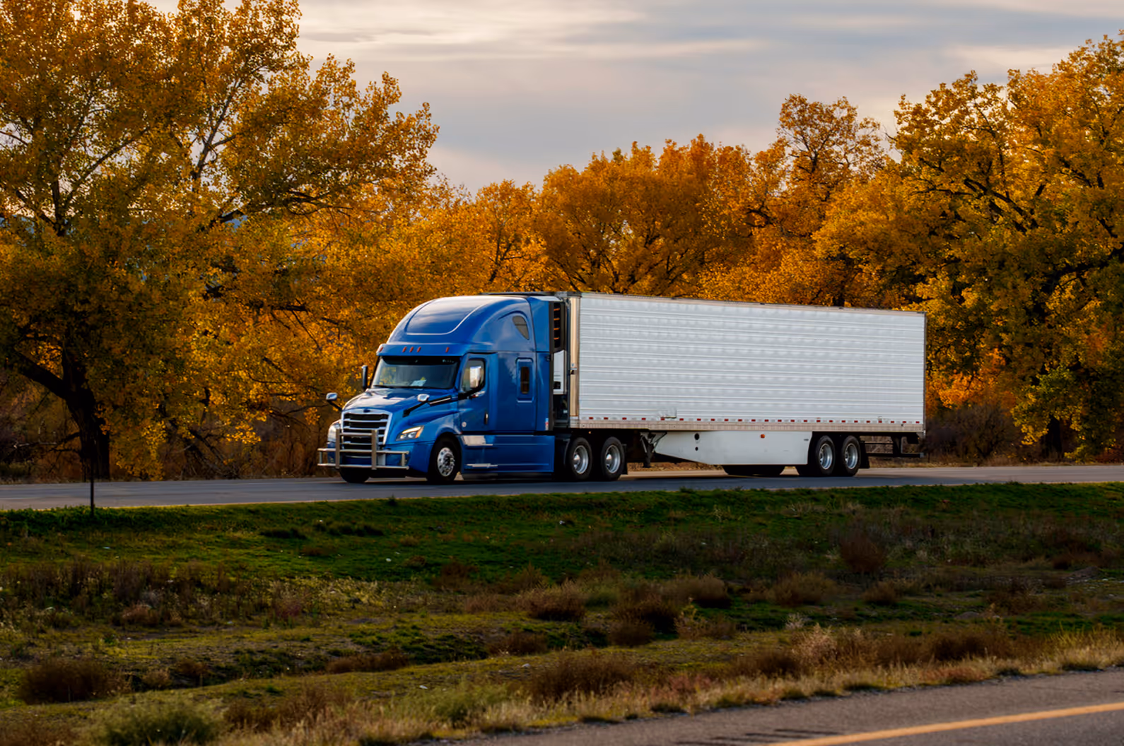 Blue semi-truck with white trailer driving on road with autumn trees in the background.