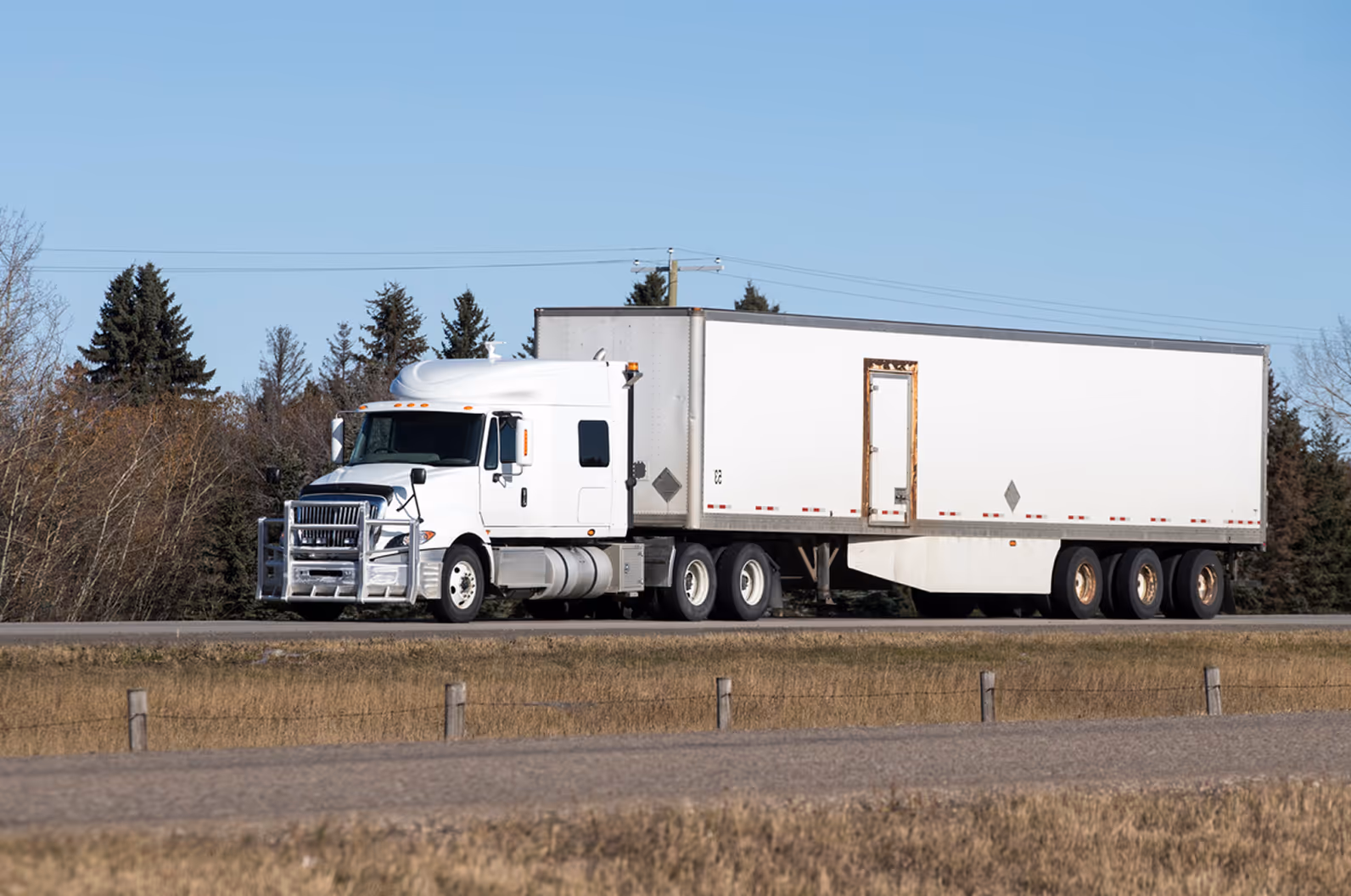 White semi-truck with a large white trailer driving on a highway with trees in the background.