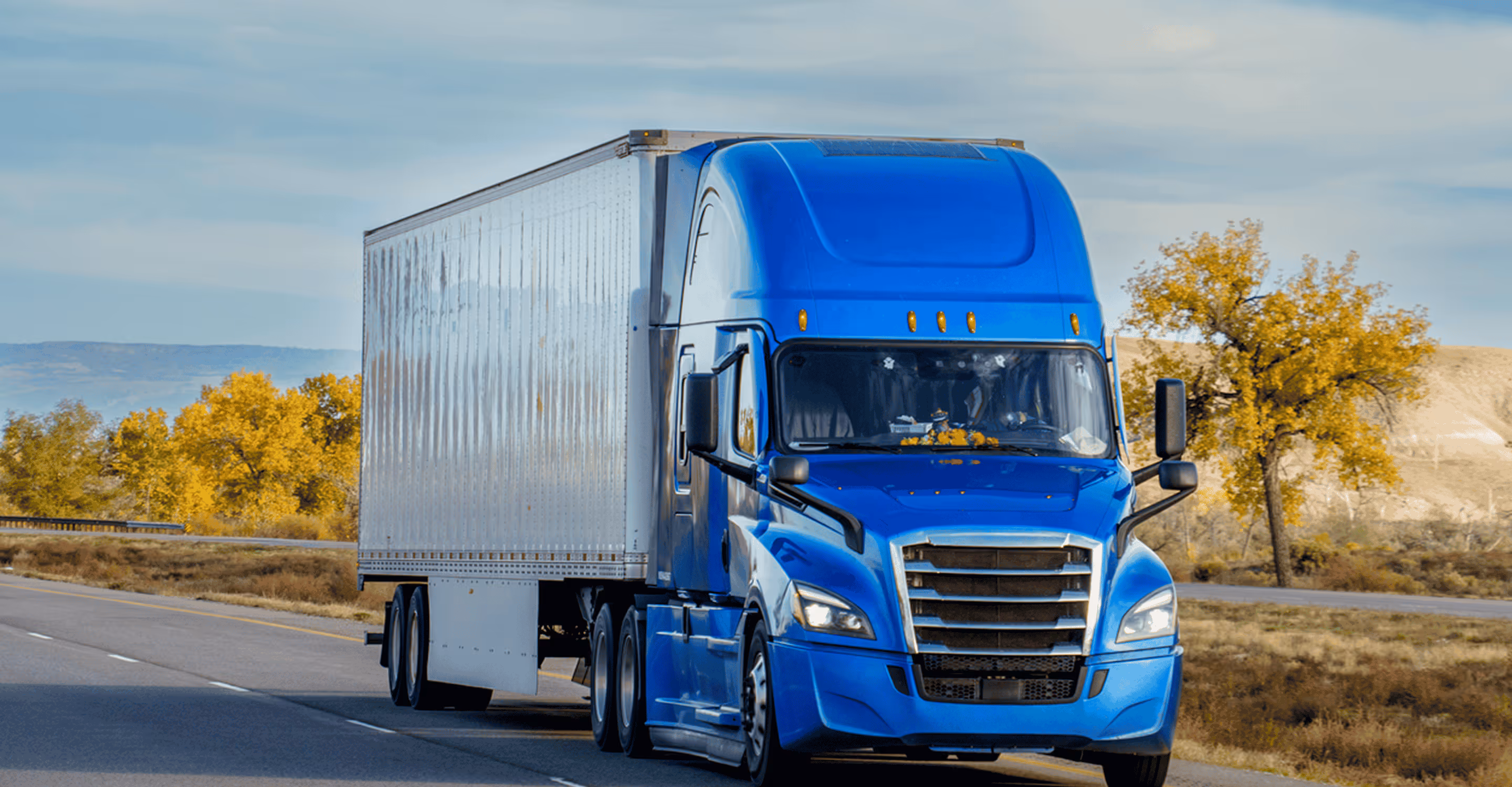 Blue semi-truck with white trailer driving on a highway with autumn trees in the background.