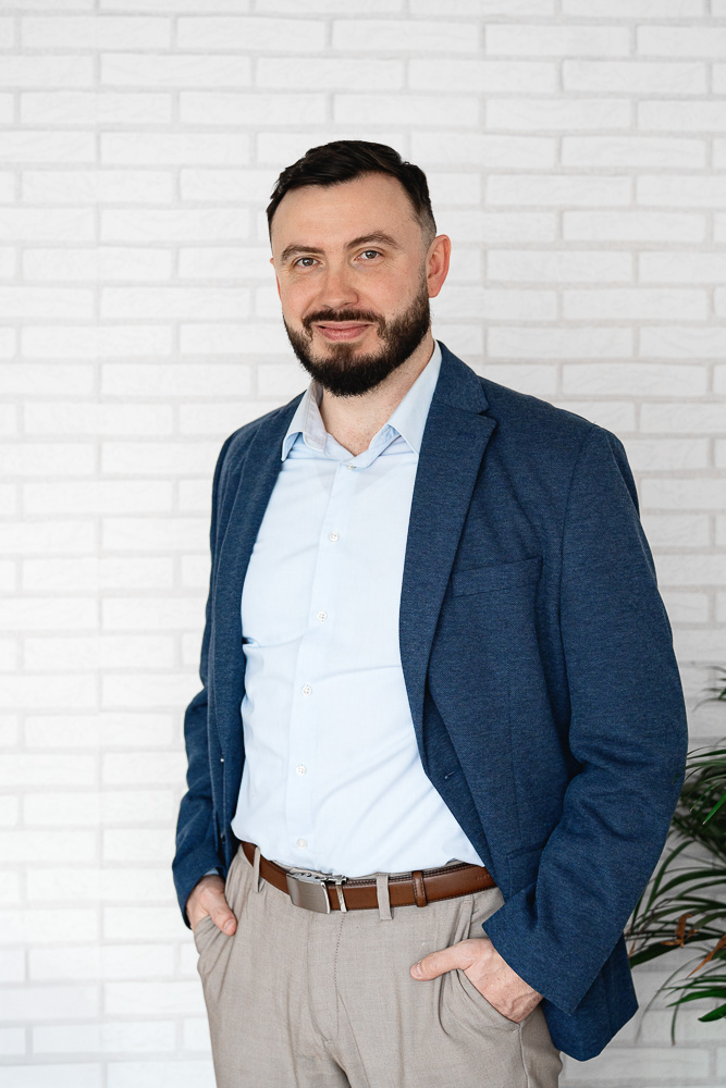 Muž s tmavými vlasy a vousy v modrém tričku a tmavě modrém saku před modrým pozadím.