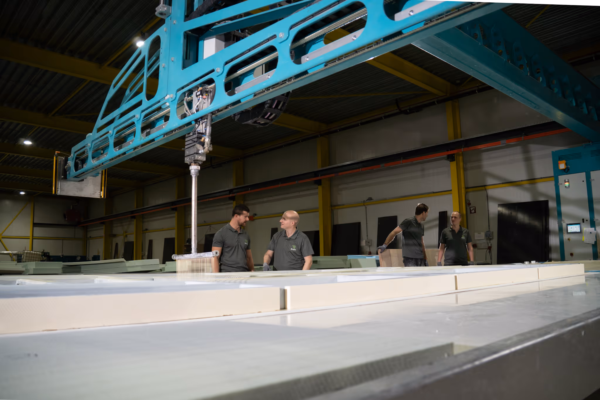 Four factory workers standing near large machinery in an industrial warehouse setting.