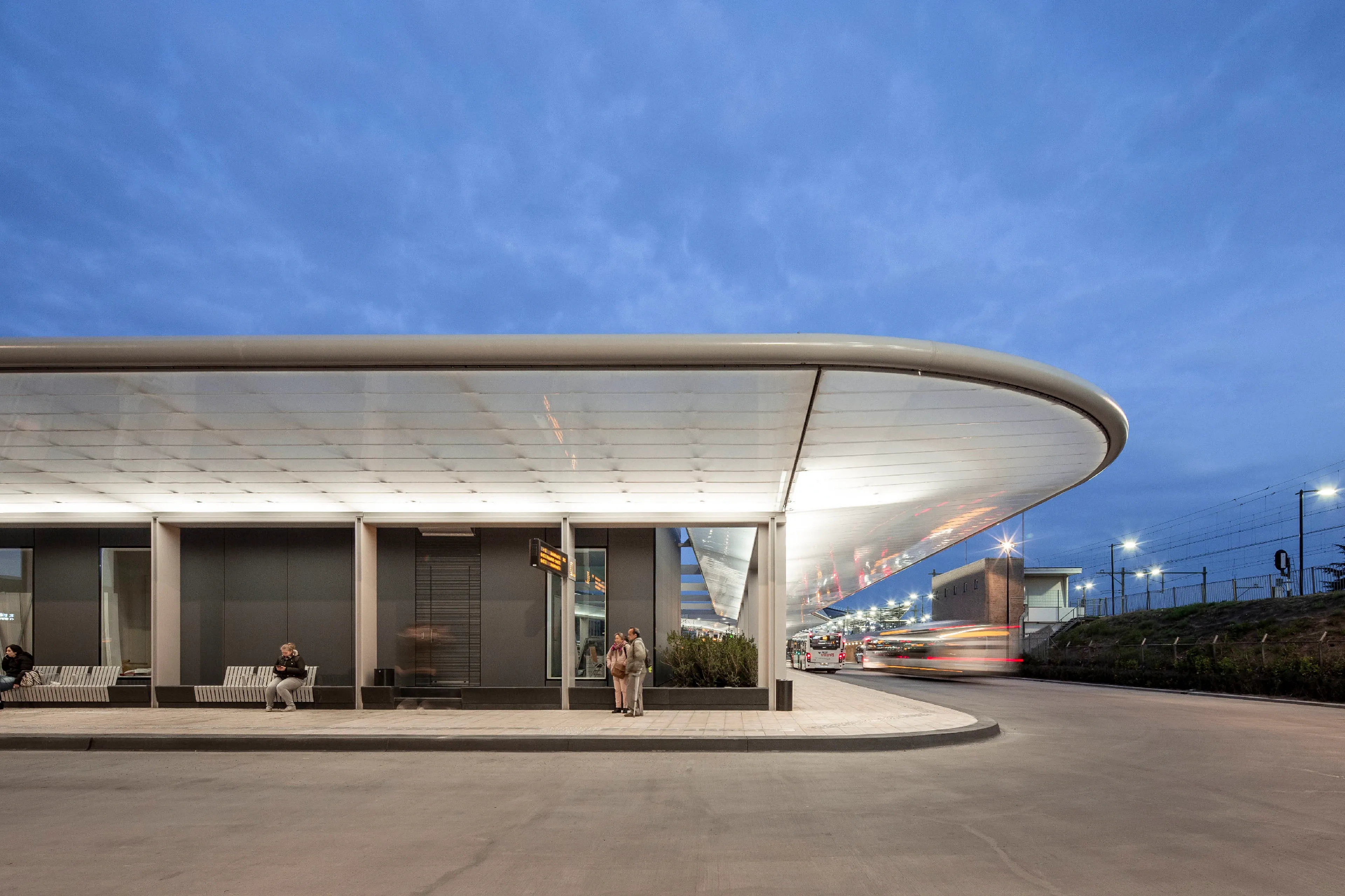Modern bus station at dusk with curved roof, people waiting, and buses passing in motion blur.