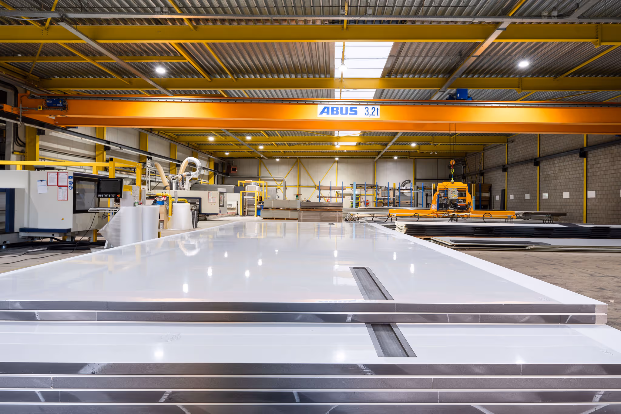 Large white metal sheets stacked in a factory with yellow cranes and industrial machinery.