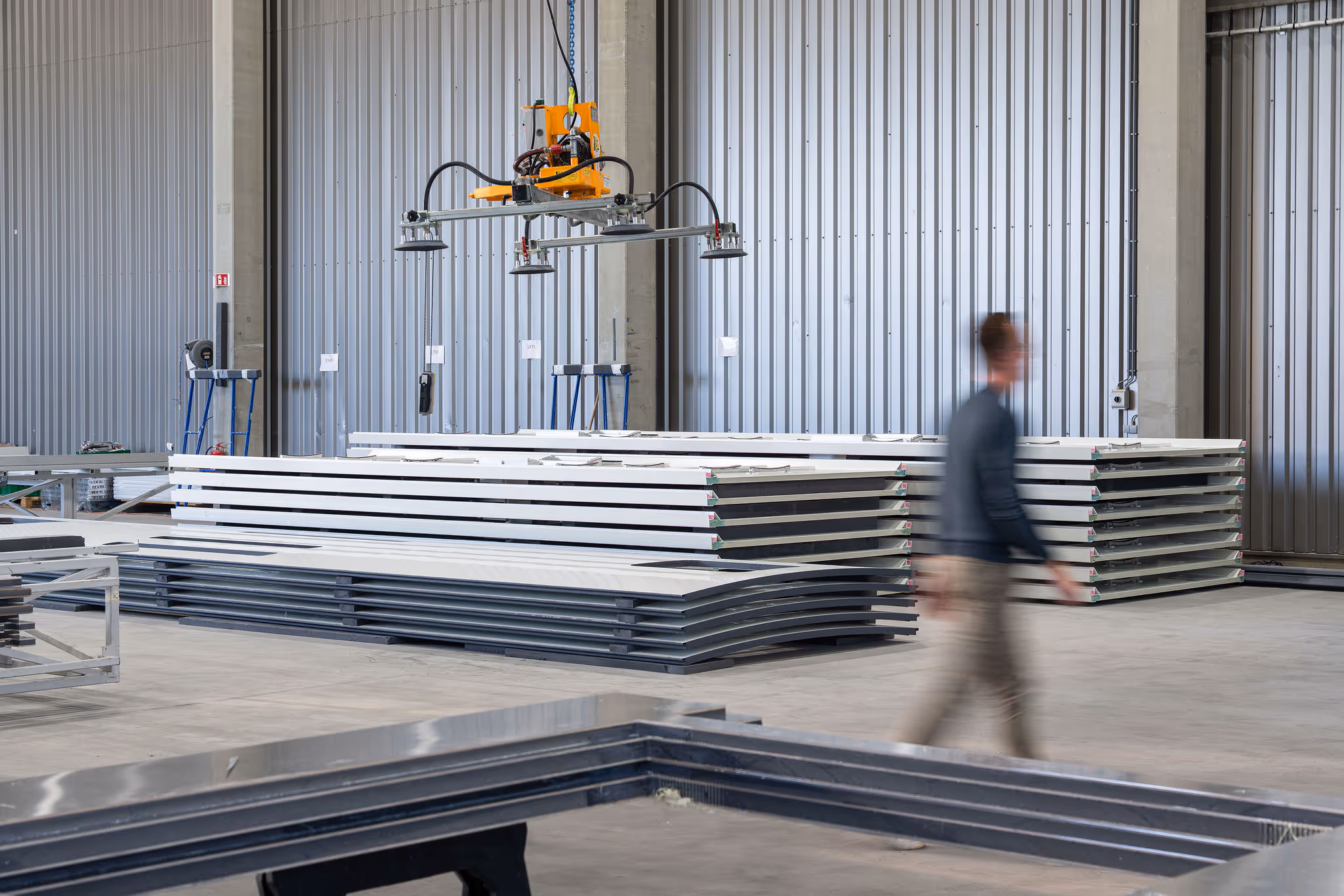 Blurred man walking in warehouse with stacked metal or panel sheets and industrial lifting equipment above.