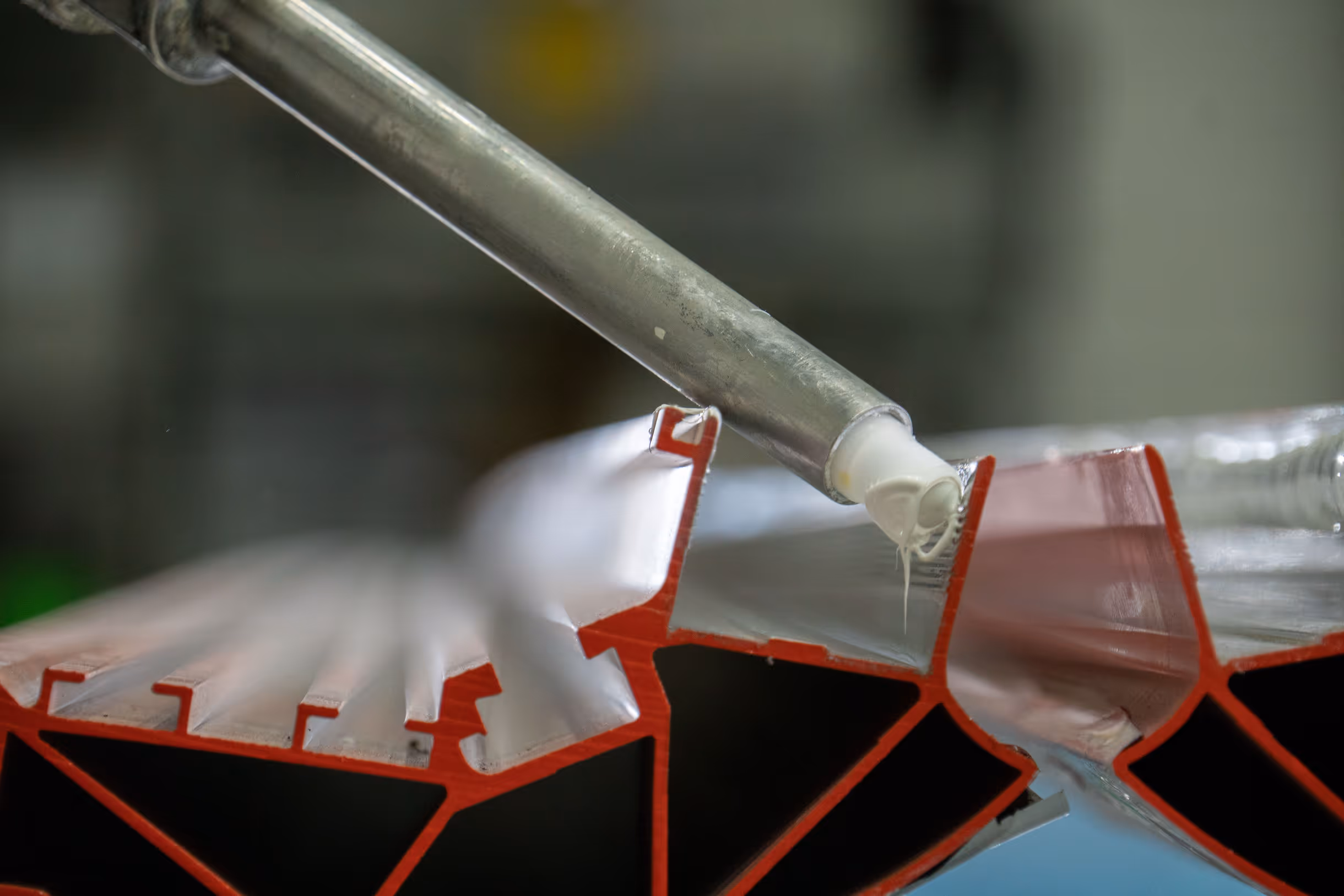 Close-up of a tool applying white adhesive to the edge of a red and silver metal extrusion.