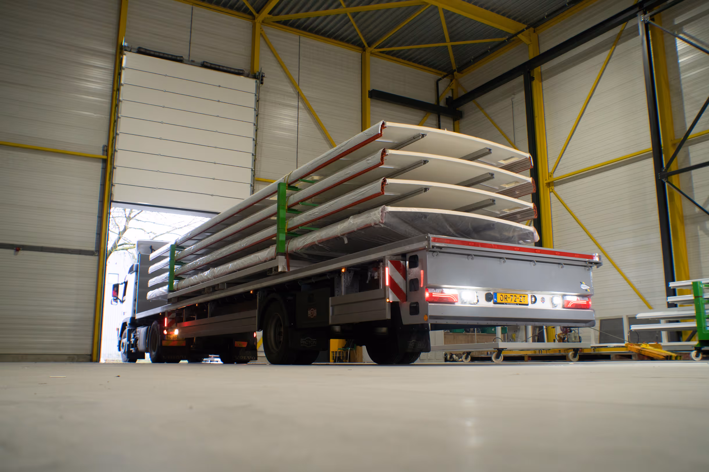 Flatbed truck loaded with long white panels inside a large industrial warehouse.