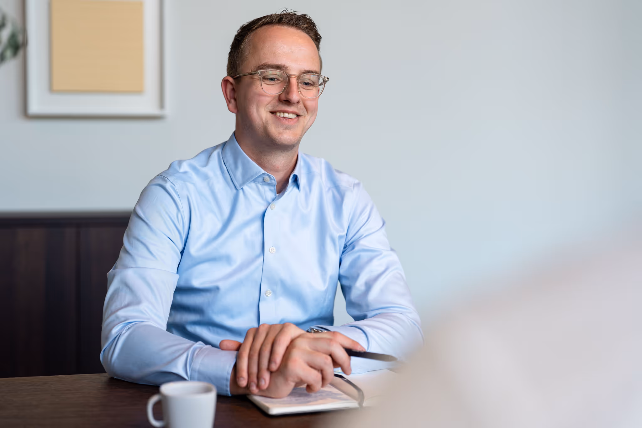 Smiling man in glasses and blue shirt sitting at a table with a notebook and coffee cup.