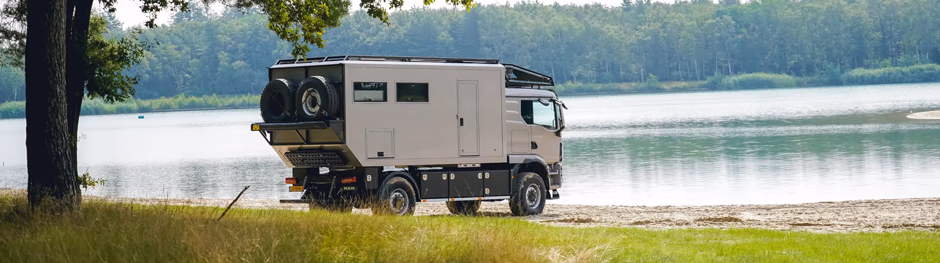 Large beige camper truck parked on a grassy shore near a calm lake with trees in the background.