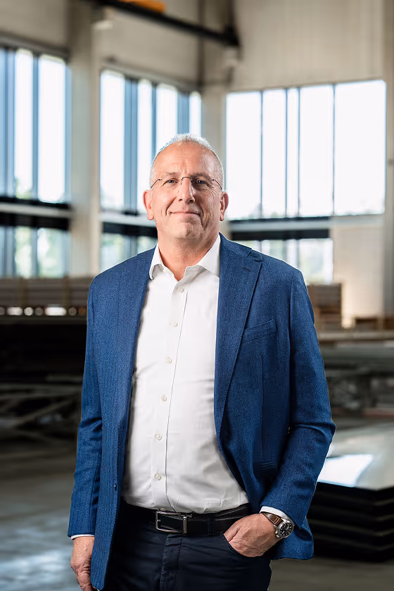 Middle-aged man in glasses and blue blazer standing in a bright industrial space with large windows.