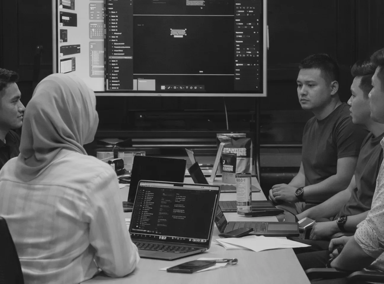 Team of young professionals having a meeting with laptops open and a large screen displaying software interface in a conference room.