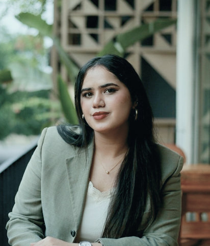 Portrait of Ayesha Malik, wearing a light-colored blazer, smiling softly in a bright outdoor setting.