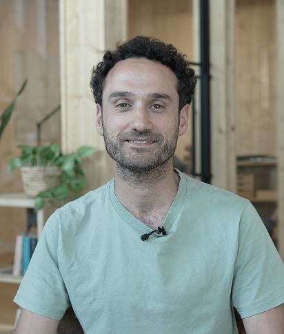 Portrait of Husam Al-Dakak, wearing a light green t-shirt, smiling in a modern wooden studio.
