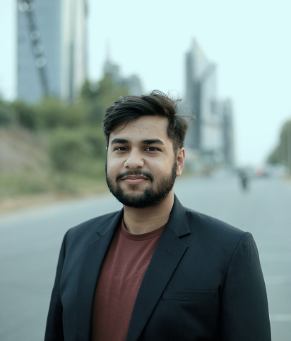 Portrait of Sher Jan, wearing a dark blazer and a maroon shirt, with a blurred cityscape in the background.