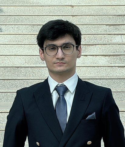 Portrait of Farhat Abbas, wearing glasses and a dark suit with a tie, against a neutral brick wall background.