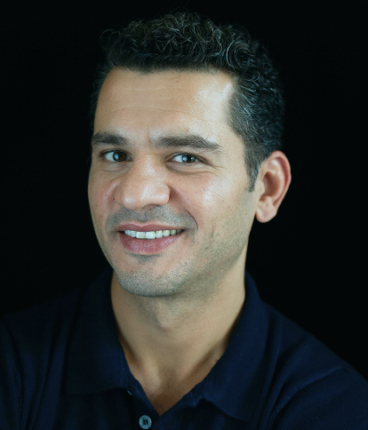 Portrait of Kais Bitar, smiling, wearing a dark polo shirt against a dark background.