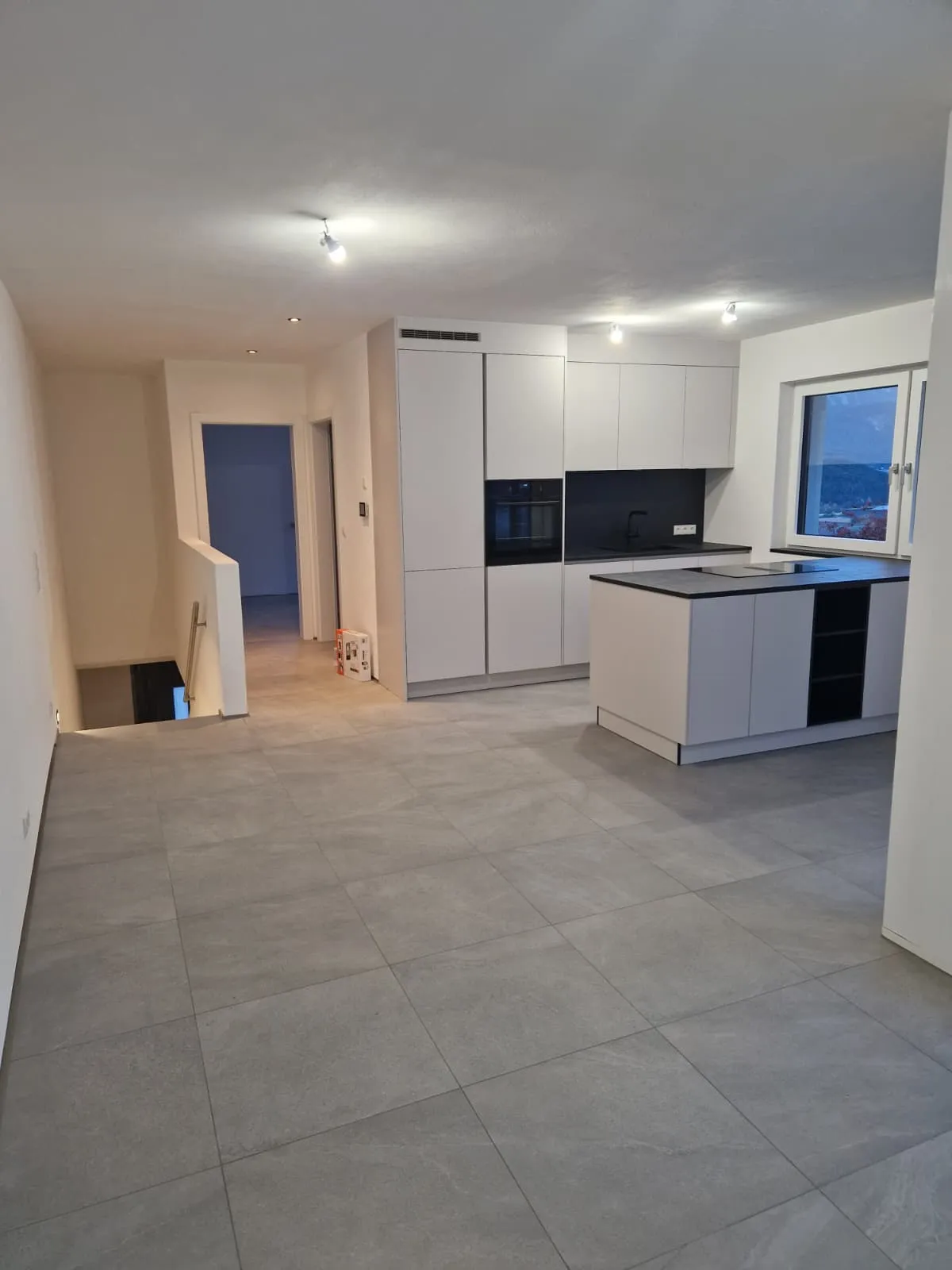 Moderne helle Küche mit großem Fliesenboden, weißer Einbauküche und Kücheninsel, offen zu Wohnraum mit Treppenabgang.