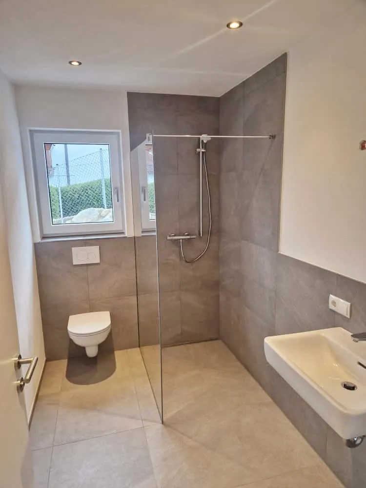 Modernes Badezimmer mit bodenebener Dusche, wandhängendem WC und weißem Waschbecken mit grauen Fliesenwänden.