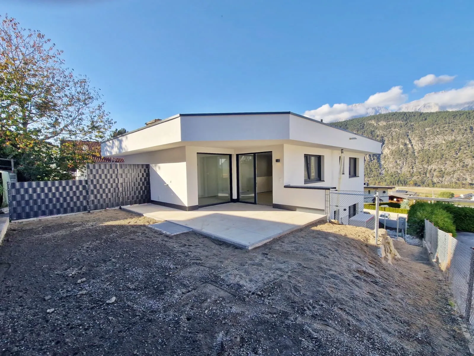 Moderner weißer Bungalow mit großer Terrassenfläche vor einem bewachsenen Hang und Bergen im Hintergrund.
