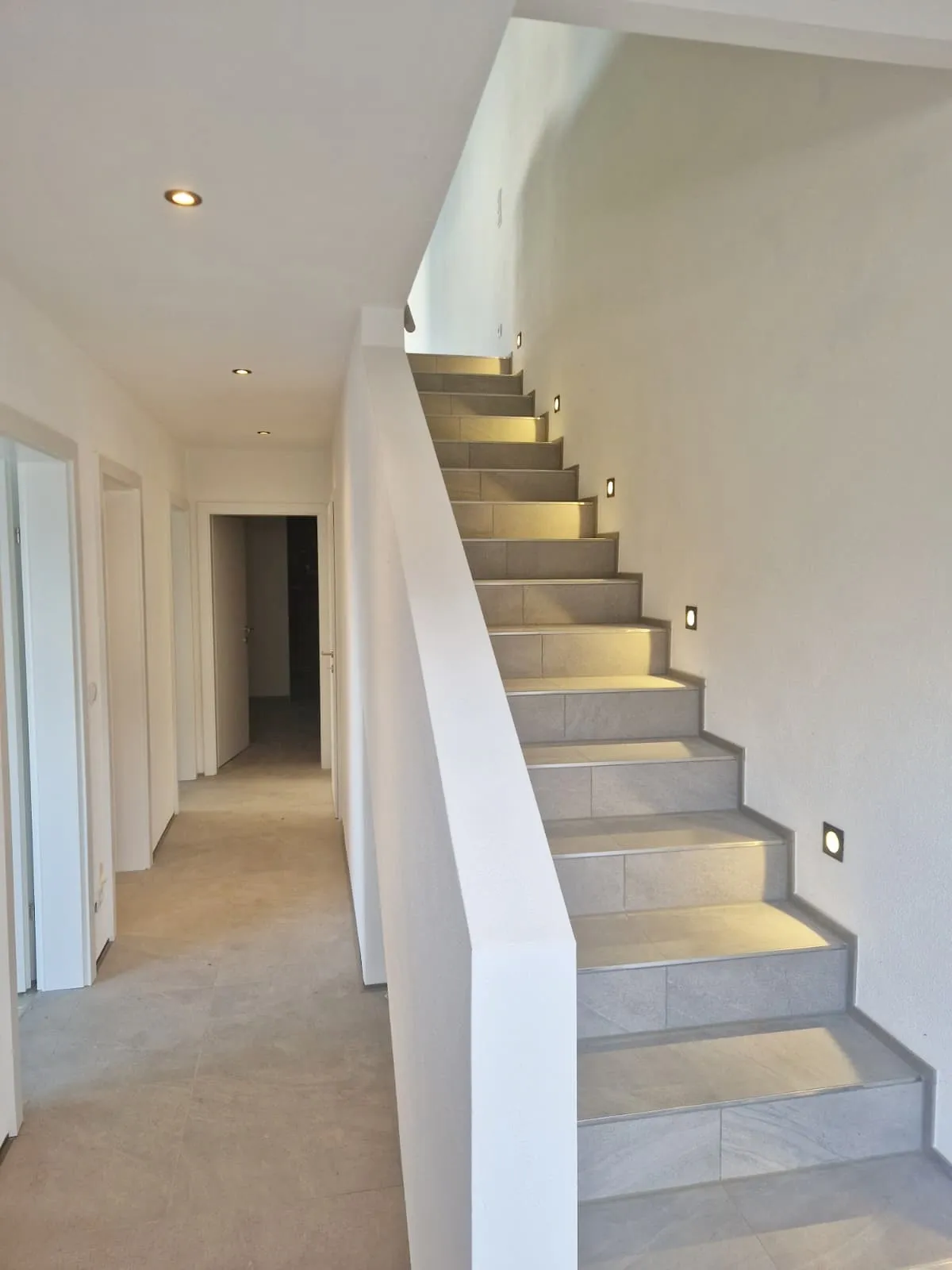 Moderner heller Innenraum mit einer Treppe aus grauen Fliesen und eingebauten Wandlichtern entlang der Stufen.
