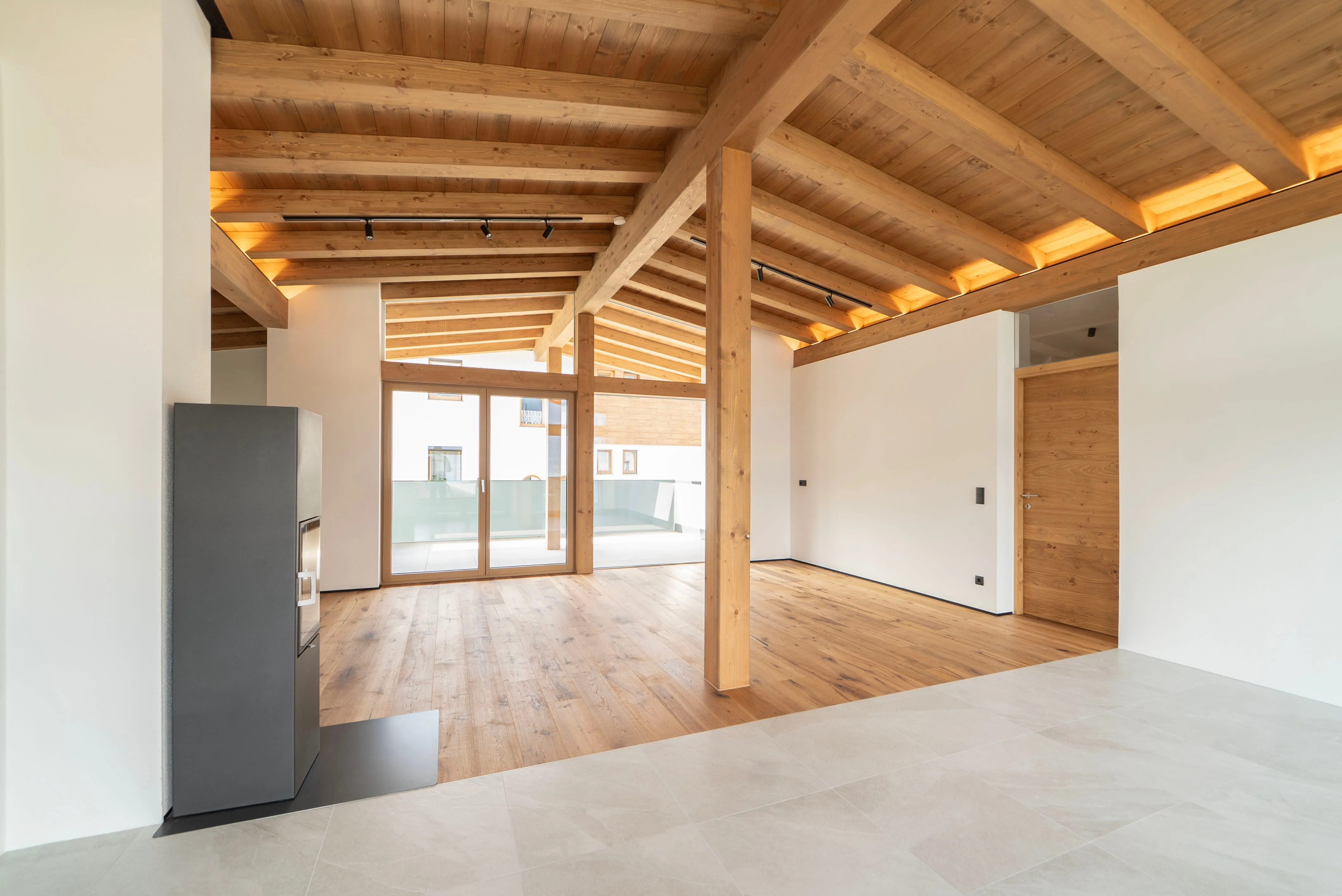 Heller, leerer Raum mit Holzdecke, Holzboden, großer Glastür zum Balkon und modernem Kamin links.