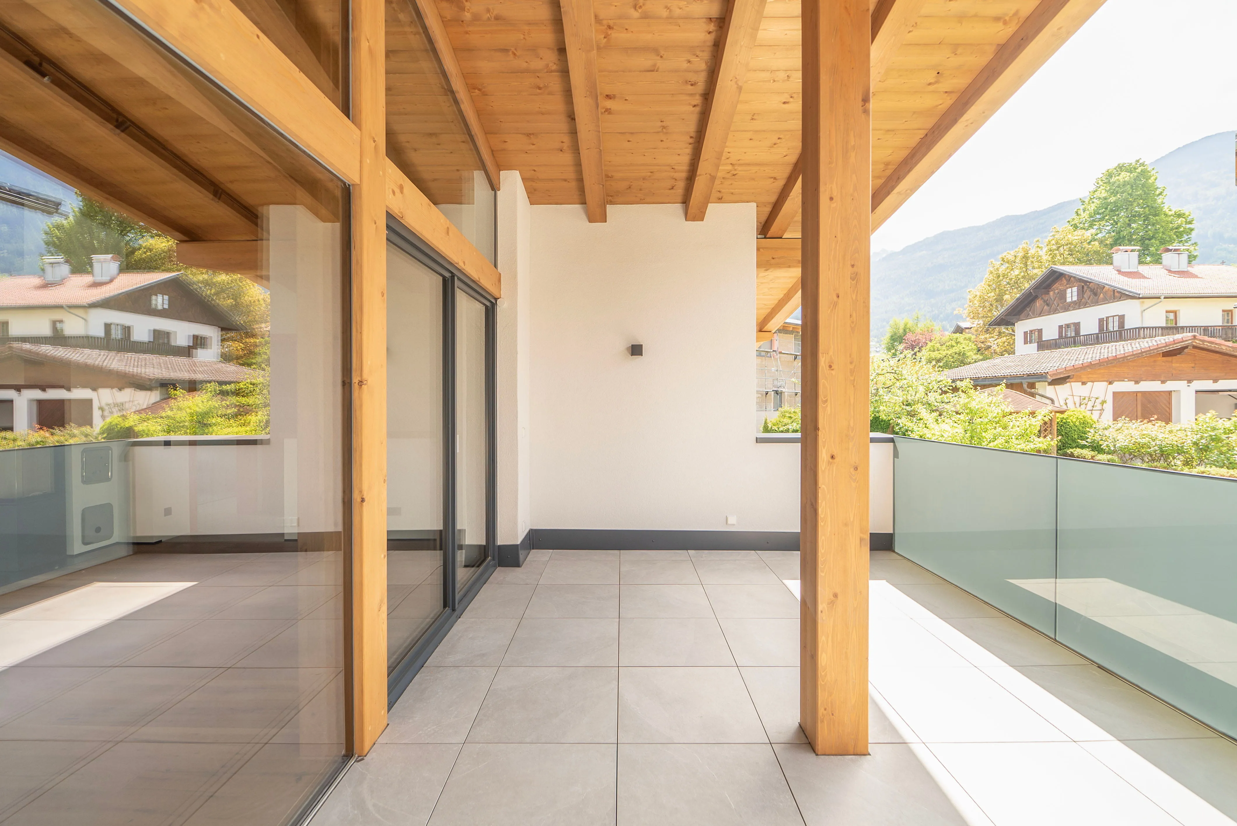 Großer, sonniger Balkon mit hellen Fliesen, Holzdecke und Glasgeländer, Blick auf Nachbarhäuser und grüne Bäume.