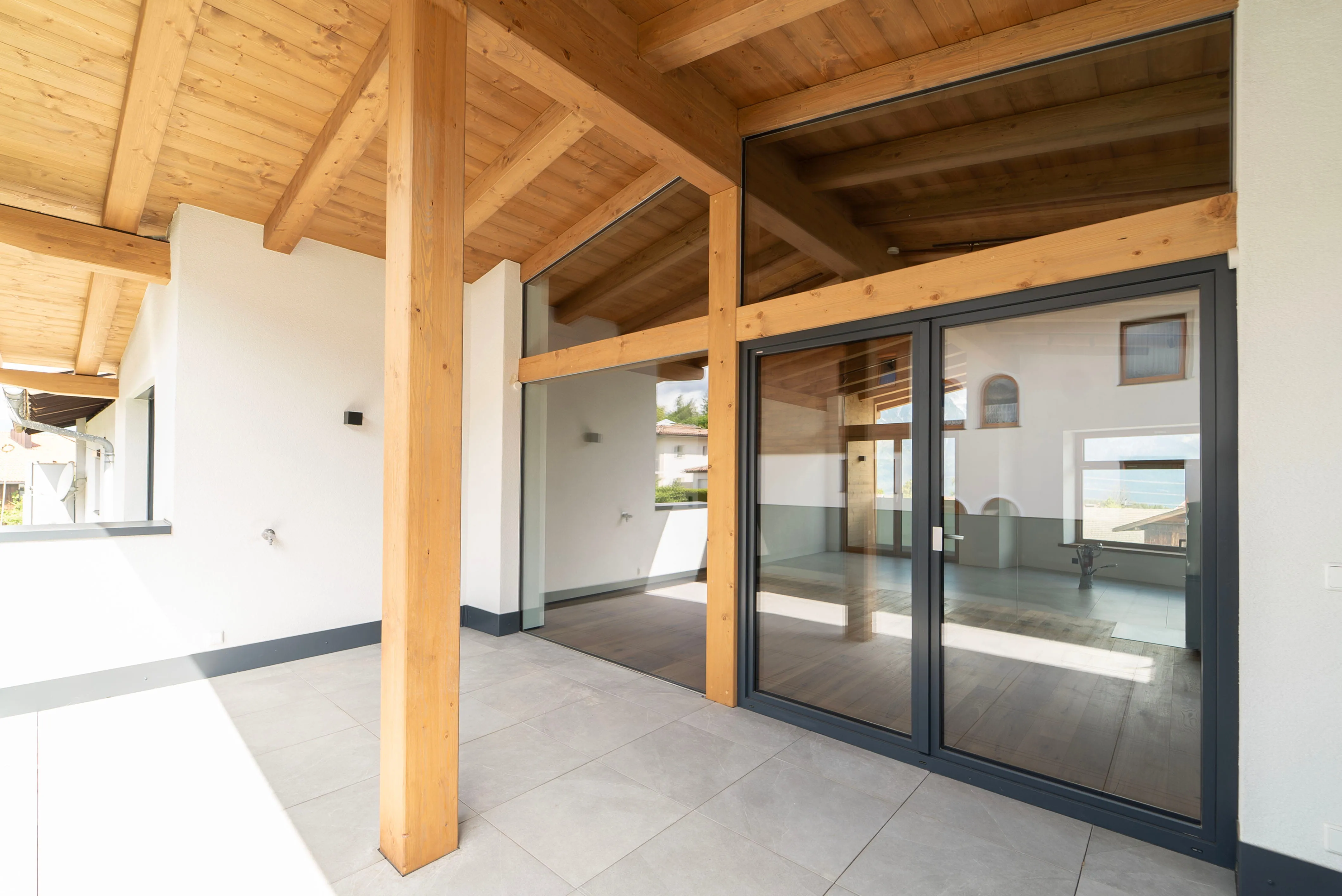 Überdachte Terrasse mit hellen Fliesenboden, Holzsäulen und großen Glas-Schiebetüren zu einem Innenraum mit Holzbalkendecke.