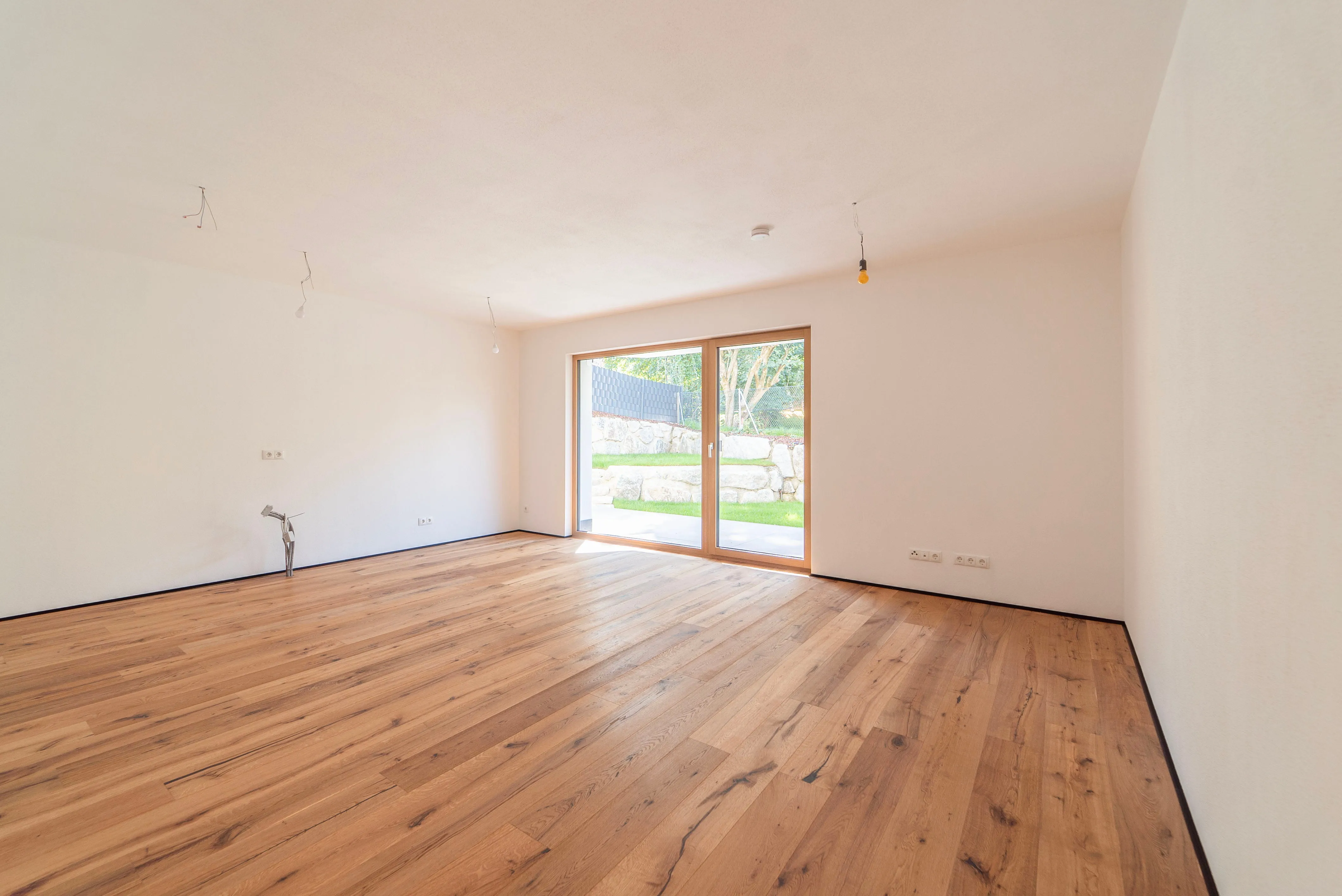 Leerer Raum mit hellen Holzböden, weißen Wänden und großen Glastüren, die auf einen Garten mit Steinen und Bäumen blicken.