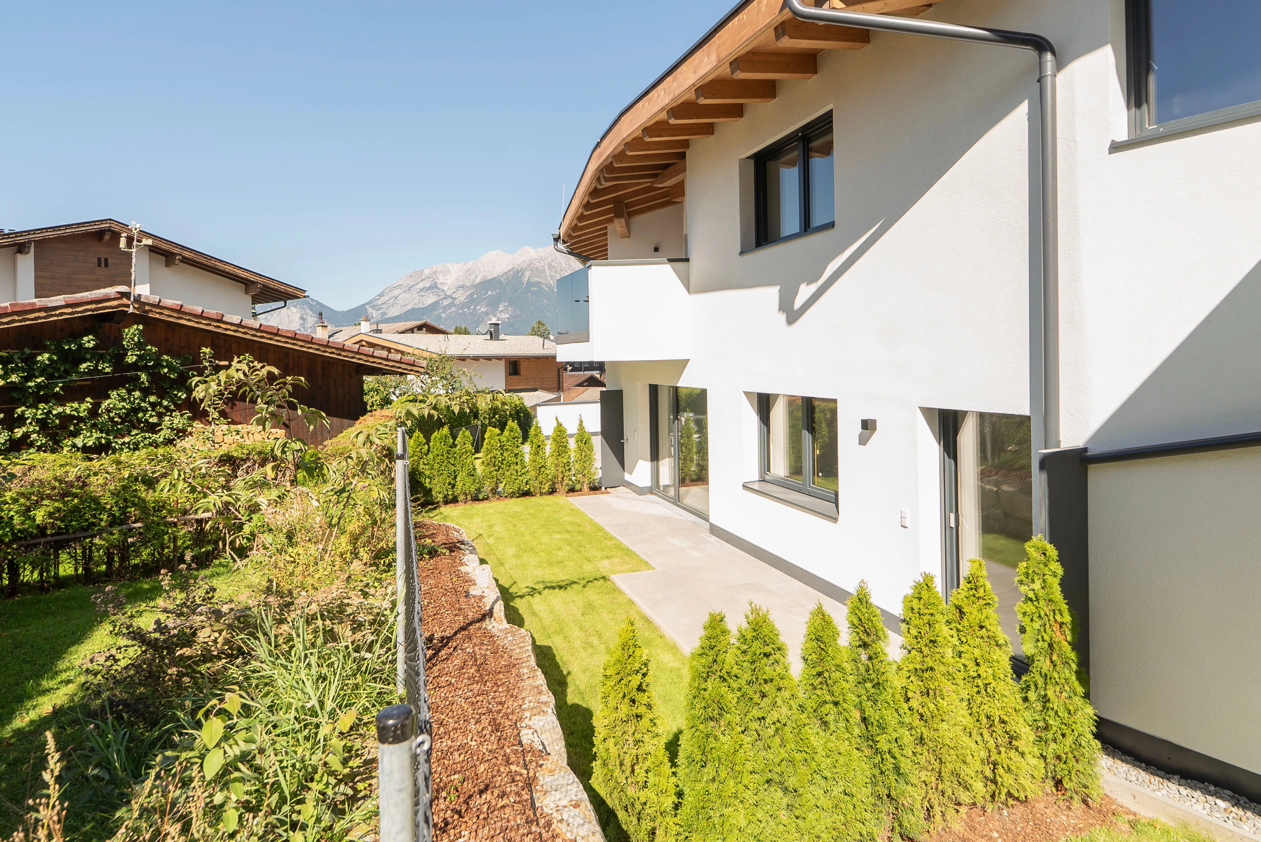 Modernes Haus mit weißer Fassade, Terrasse und gepflegtem Garten vor Bergkulisse.
