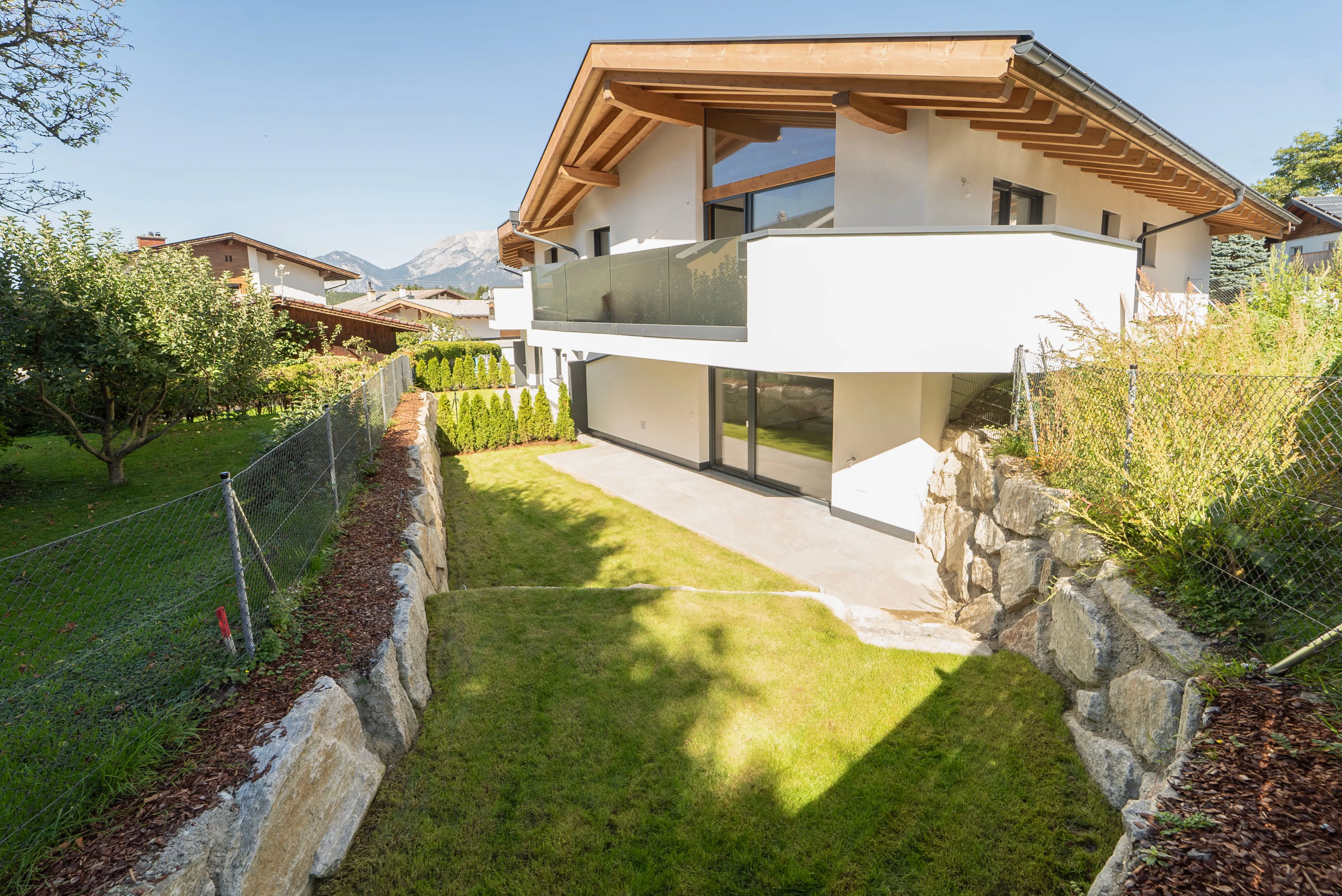 Modernes Wohnhaus mit Holzgiebel und Balkon, umgeben von einem gepflegten Garten mit Rasen und Steineinfassung.