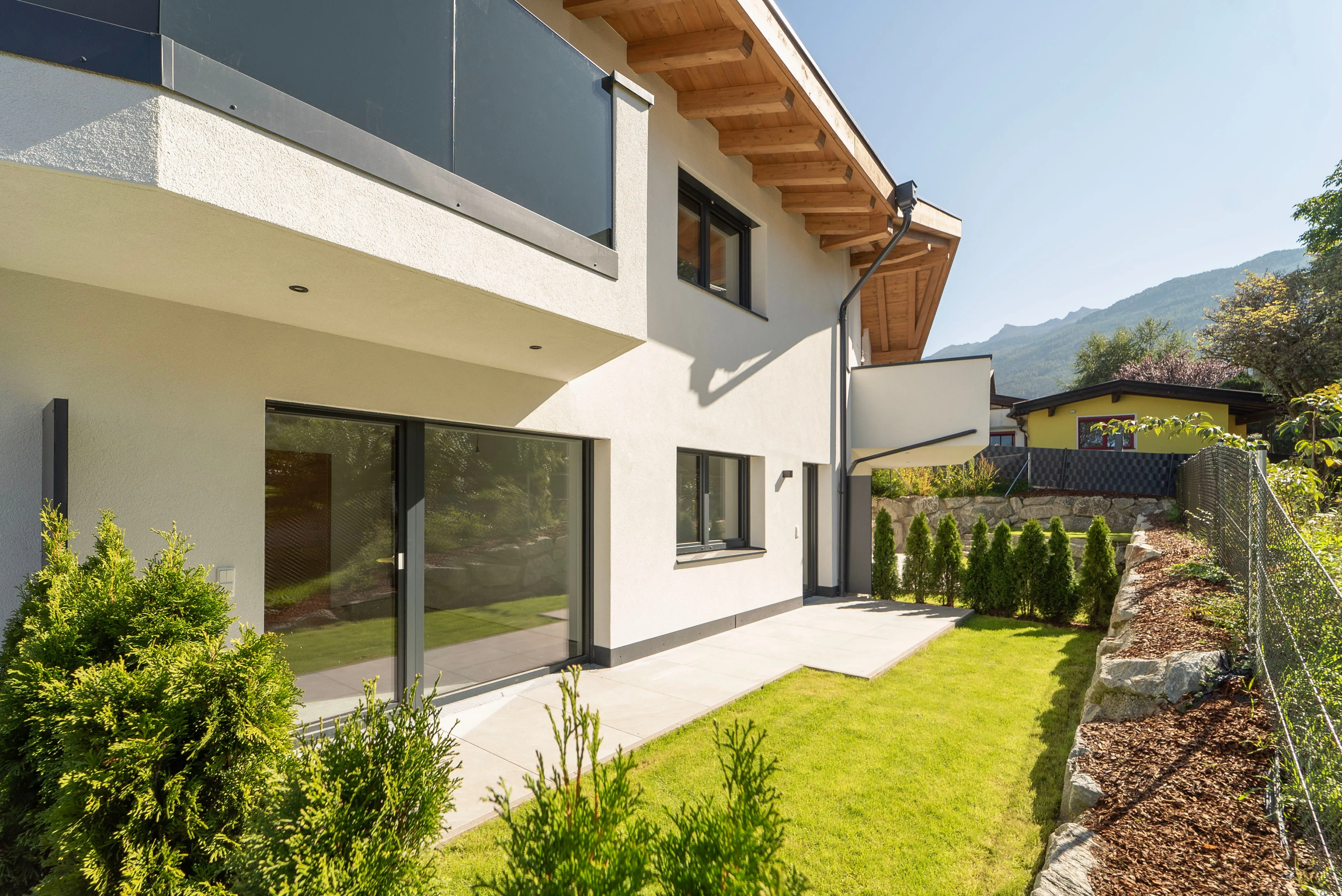 Modernes Haus mit weißer Fassade, großen Fenstern, gepflegtem Garten und gemauerter Terrasse bei sonnigem Wetter.