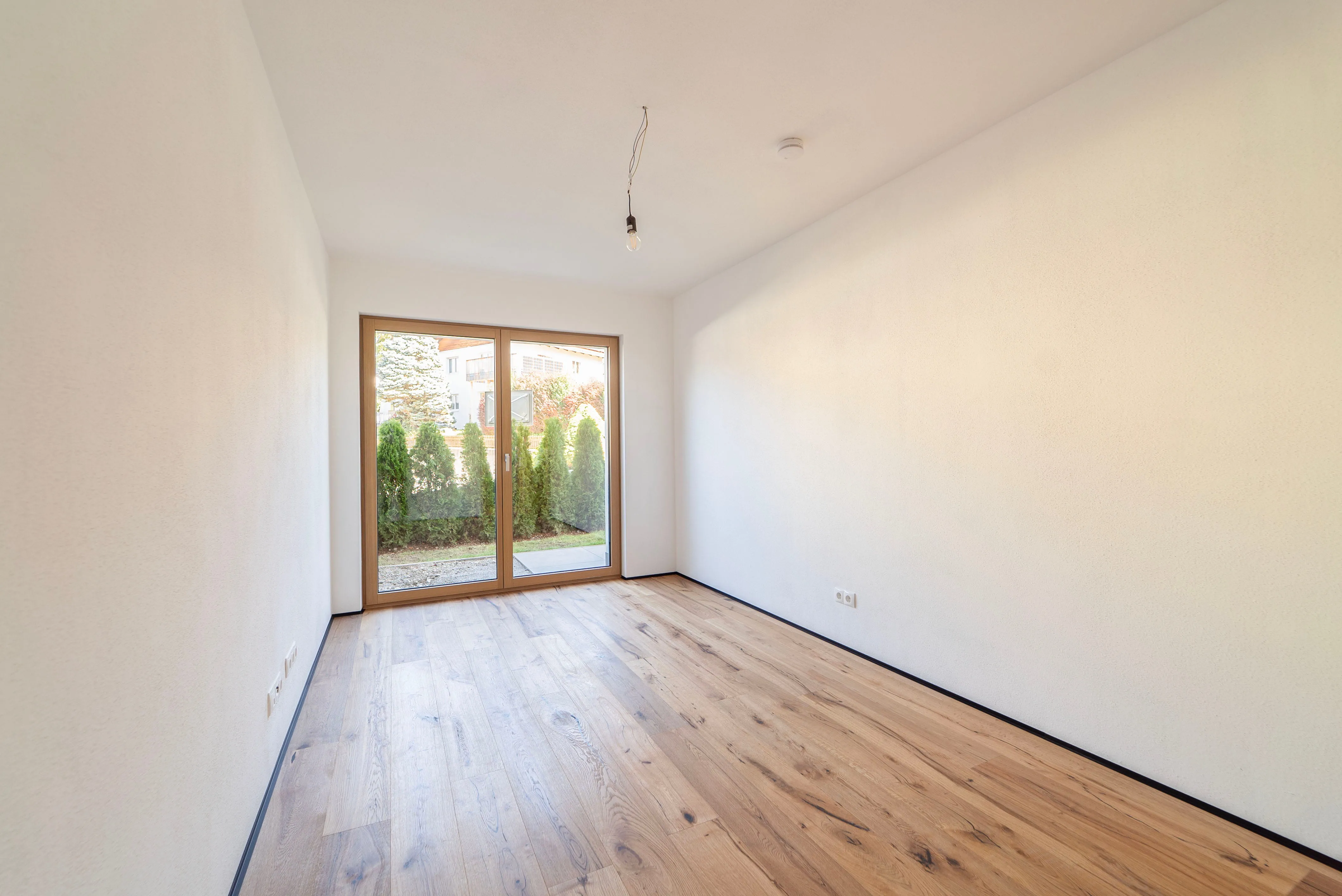 Helles, leeres Zimmer mit Holzboden und großer Glasschiebetür mit Blick auf einen Garten mit grünen Büschen.