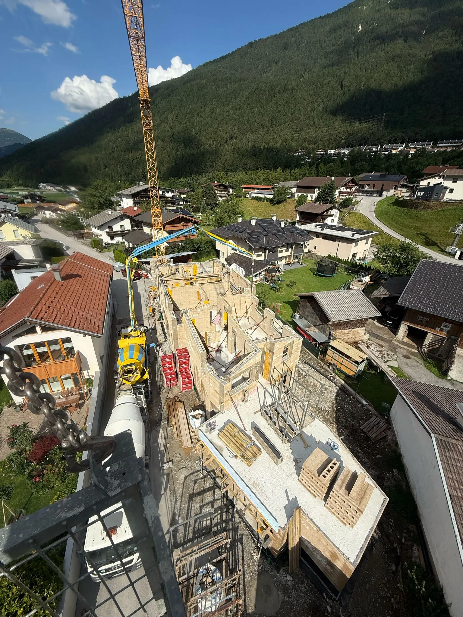 Baustelle eines mehrstöckigen Gebäudes in einem Bergdorf mit einem gelben Kran und Betonmischer, umgeben von Wohnhäusern und bewaldeten Bergen.
