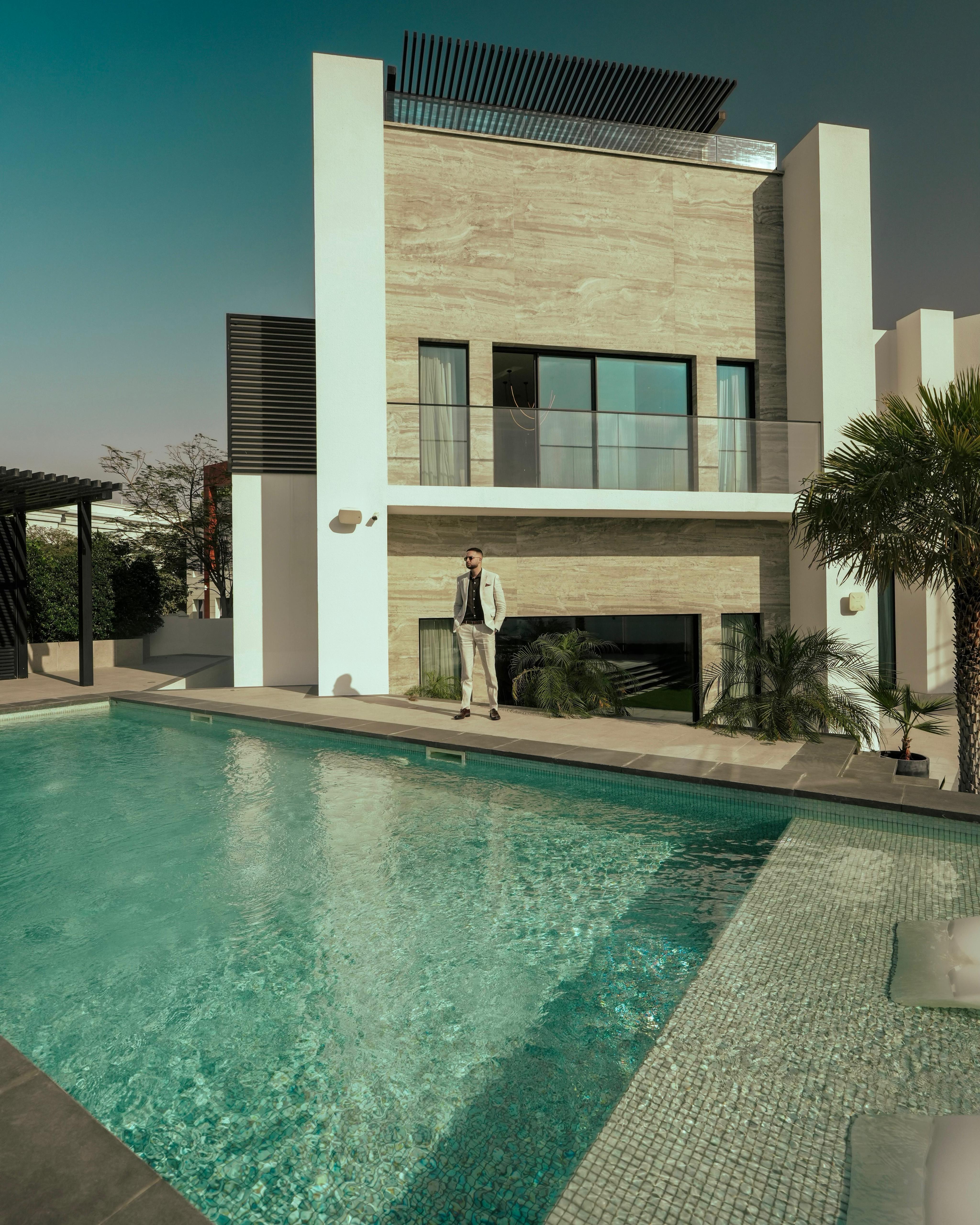 Man in a beige suit standing beside a modern house with a large swimming pool in the foreground.