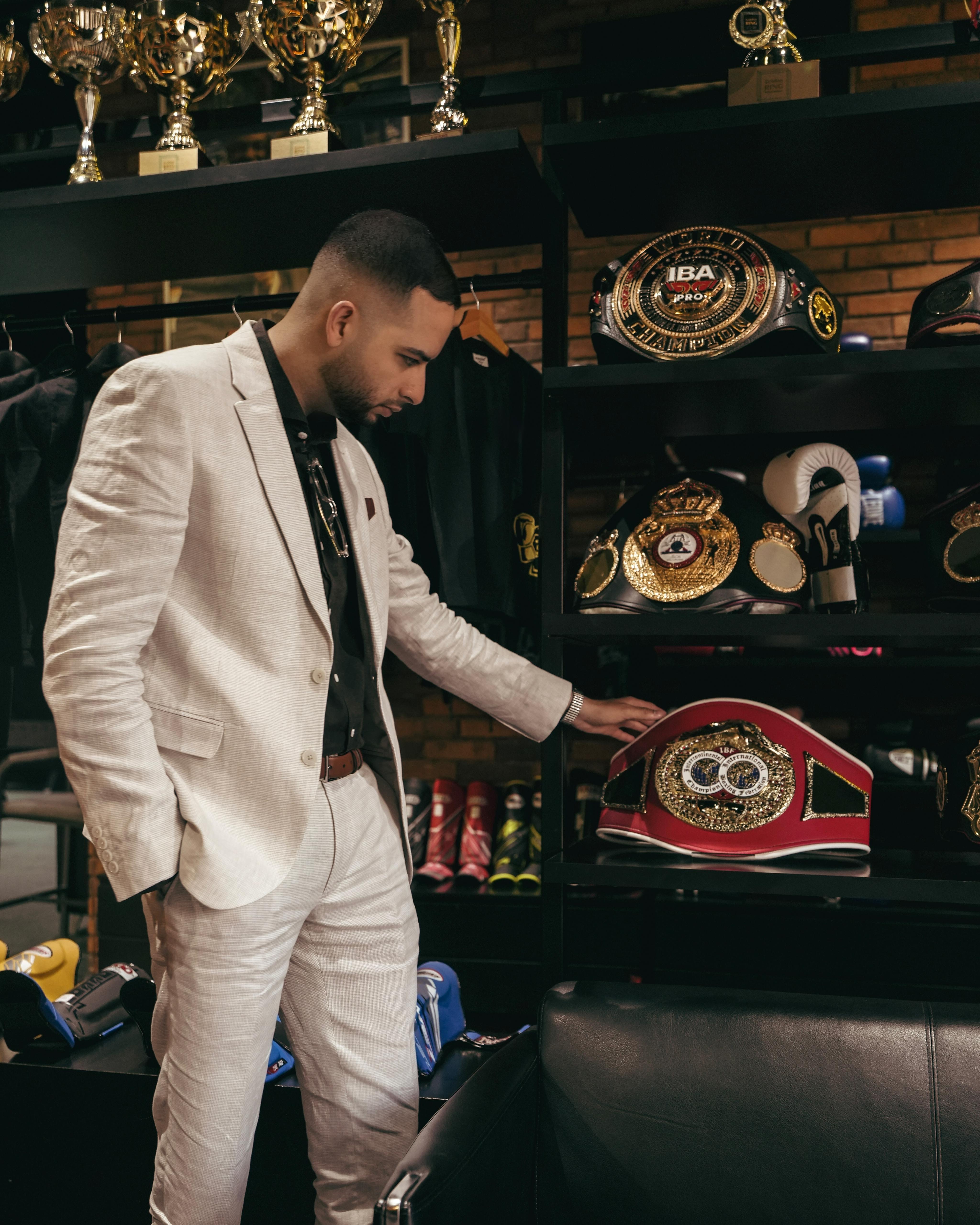 Man in a light suit looking at a collection of boxing championship belts on a black shelf.