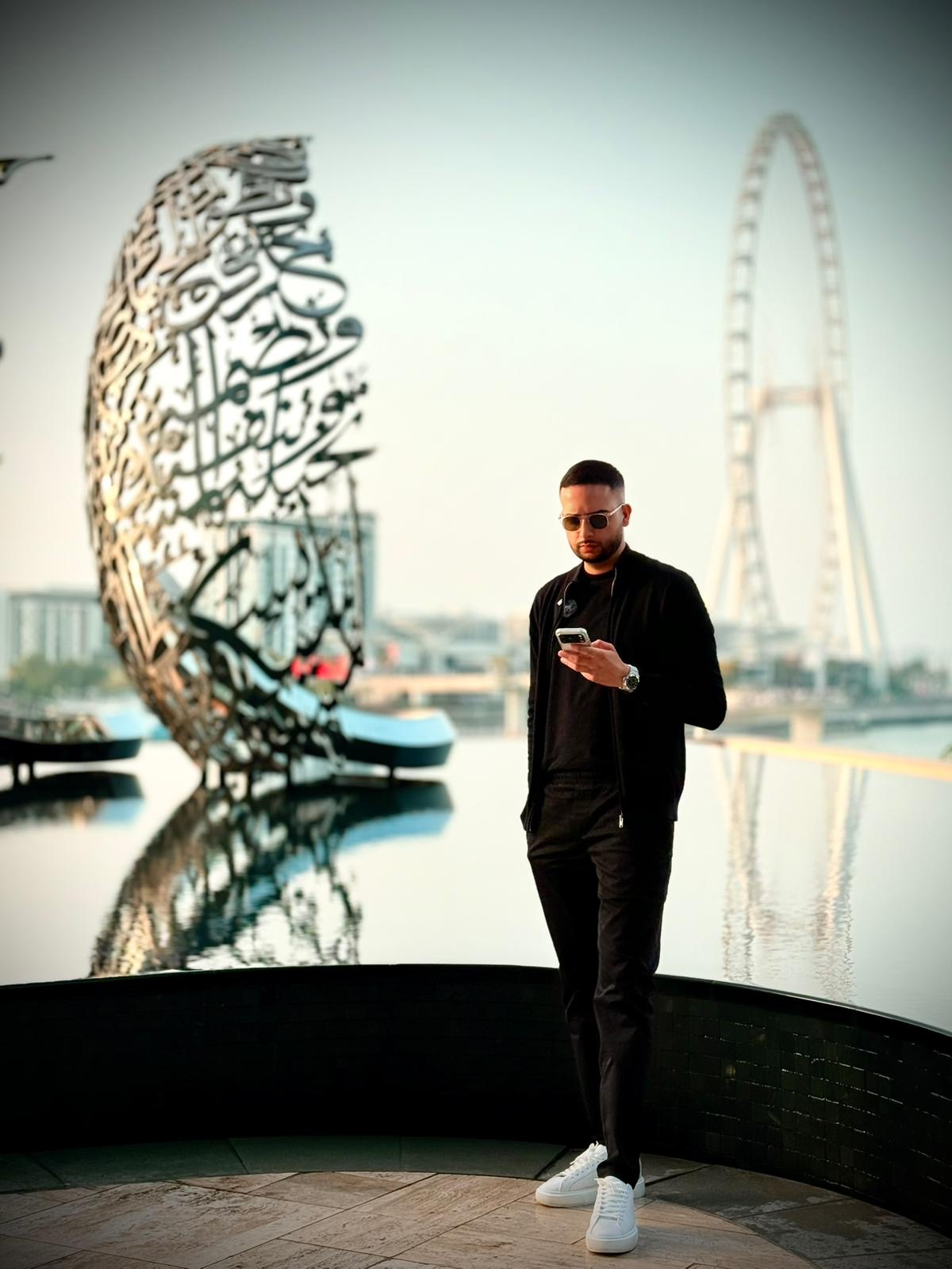 Man wearing sunglasses and black outfit standing near water with a large metal sculpture and a Ferris wheel in the background.