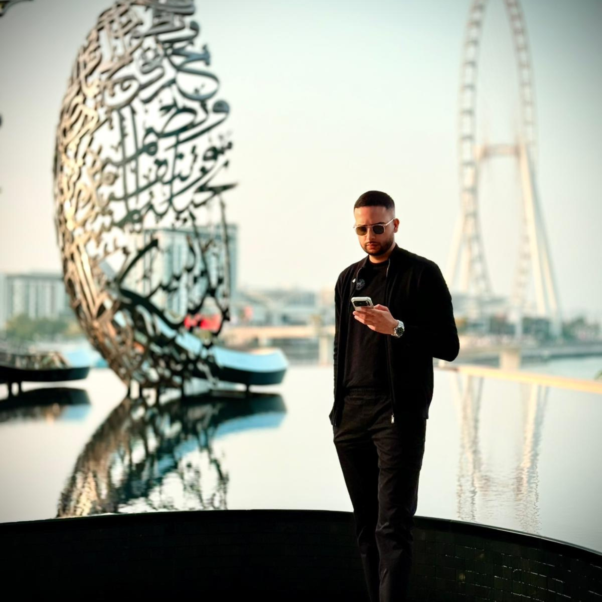 Man wearing sunglasses and black clothes looking at his phone near a reflective water surface with modern sculpture and Ferris wheel in the background.