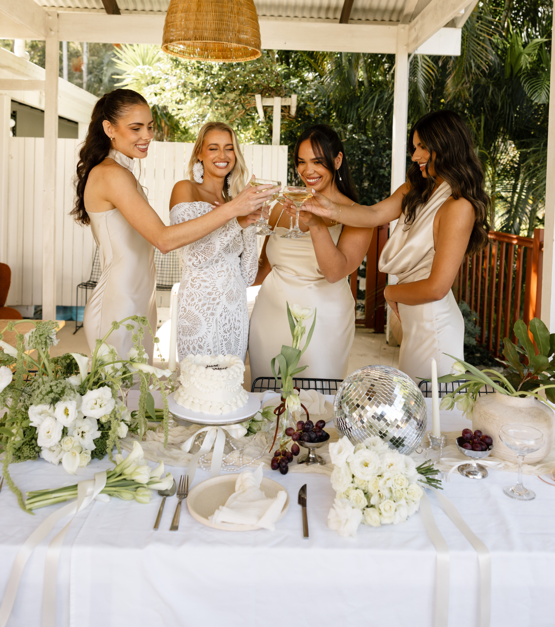 Bride and bridesmaids toasting champagne over a styled wedding table with white flowers and cake at Greenhouse