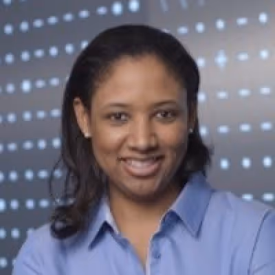 Smiling woman with dark hair wearing a light blue collared shirt in front of a blurred dotted background.