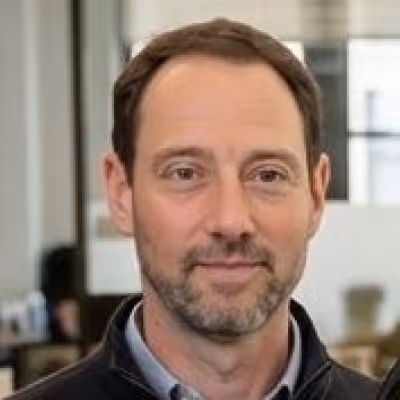 Middle-aged man with short brown hair and beard, wearing a dark jacket over a collared shirt, with an office background.