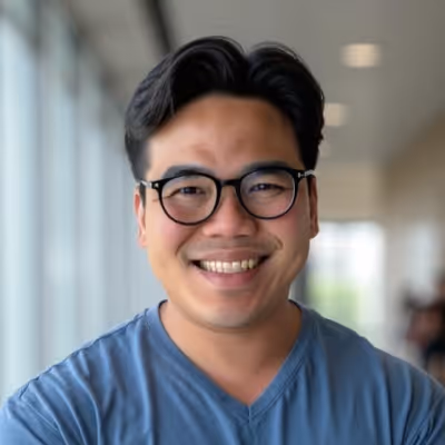 Smiling man with black hair and glasses wearing a blue V-neck shirt in a bright indoor setting.