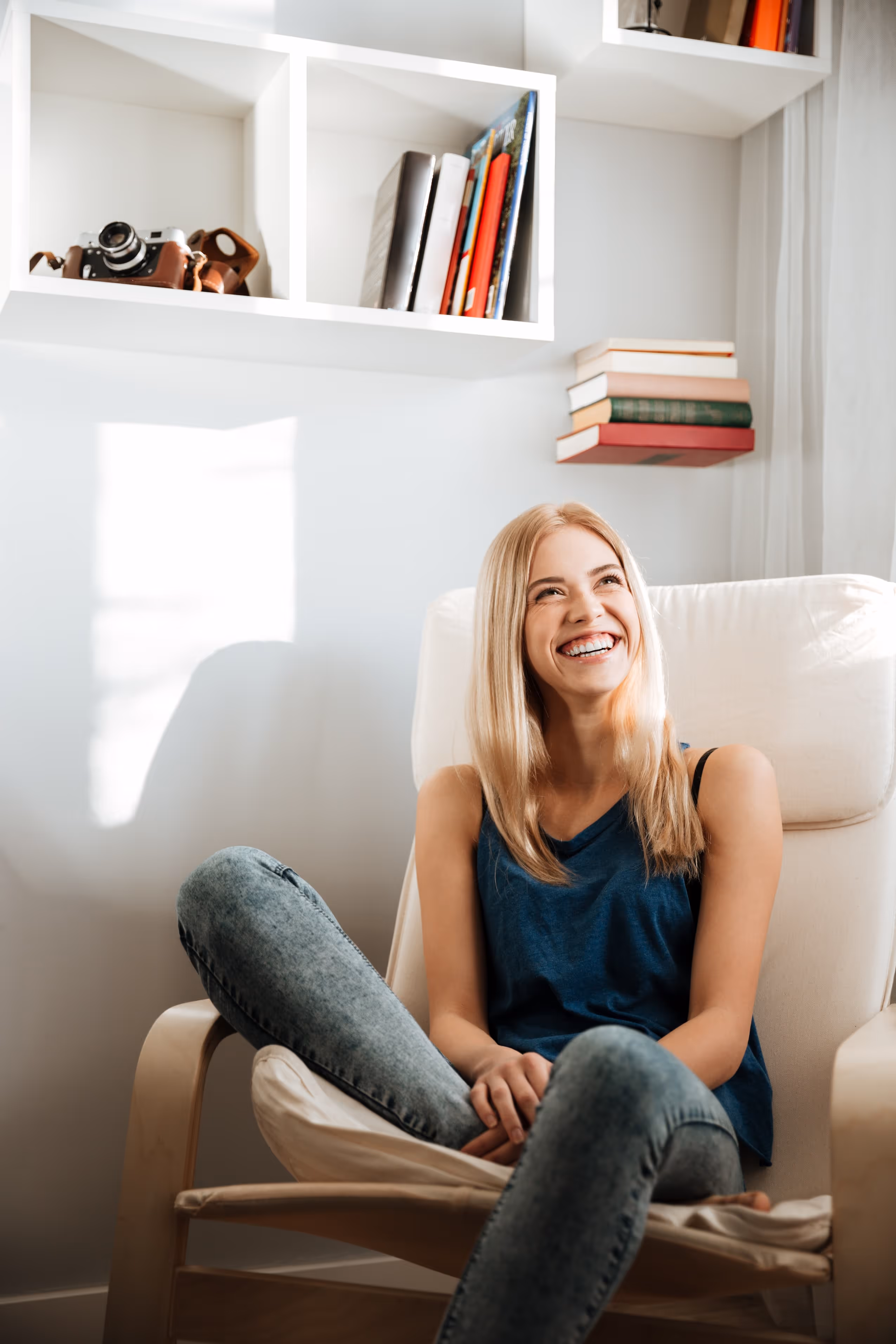 Jeune femme blonde souriante assise dans un fauteuil beige avec des étagères blanches et des livres en arrière-plan.