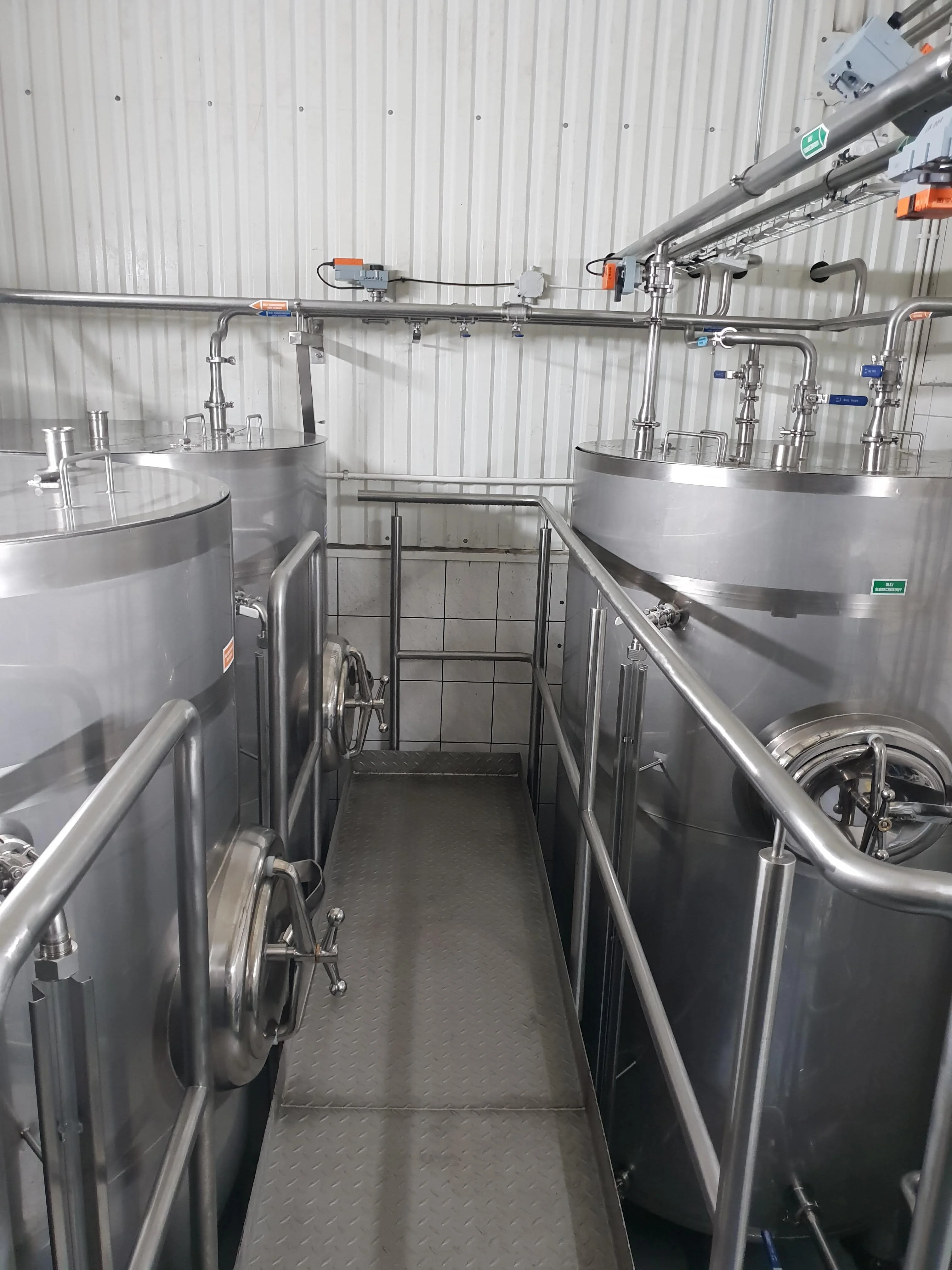 Wąski metalowy pomost między dużymi przemysłowymi stalowymi zbiornikami z zaworami i rurami w fabryce.