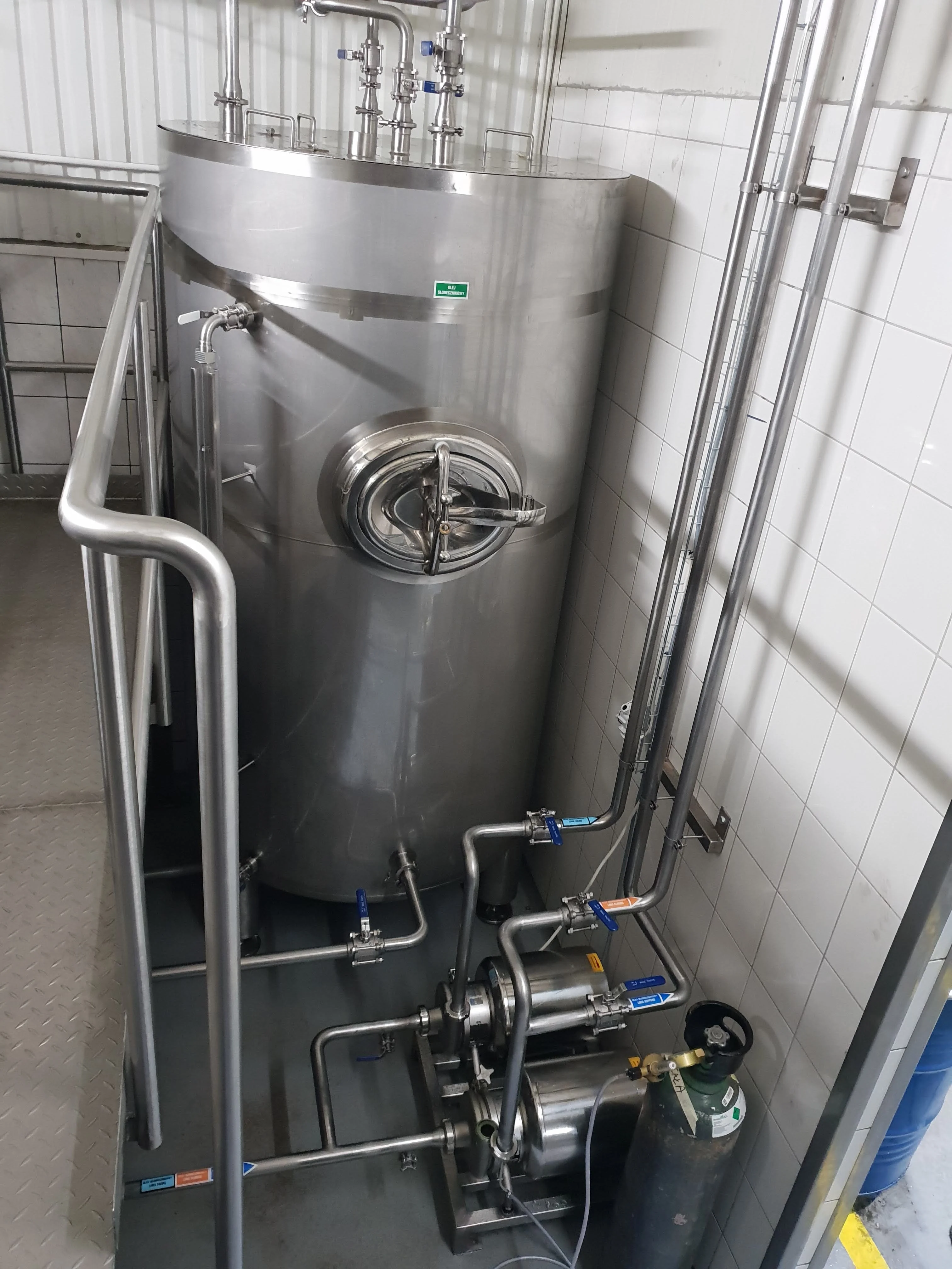 Stalowy zbiornik z rurami, zaworami i butlą gazową obok ściany w pomieszczeniu przemysłowym.