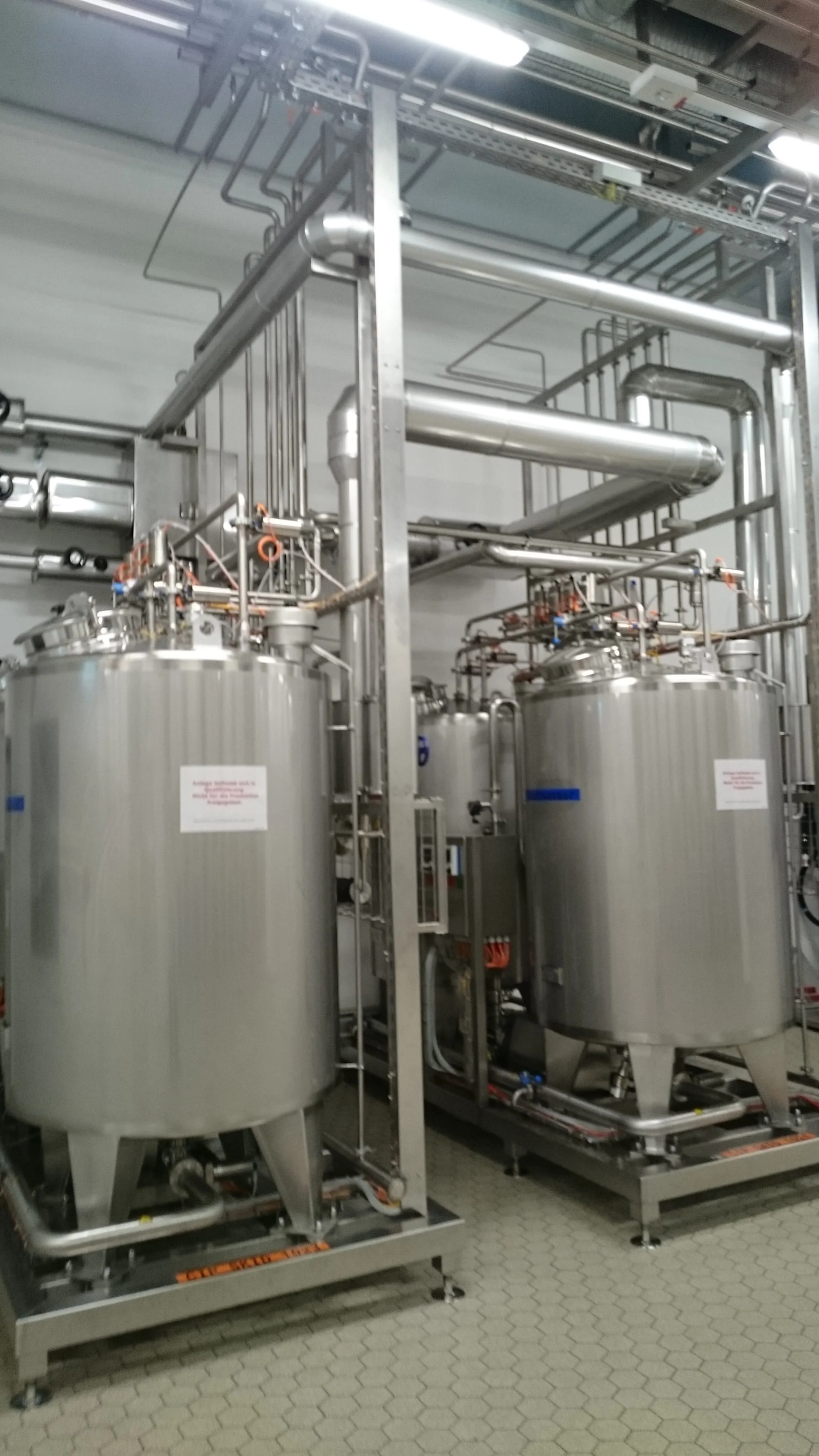 Dwa duże stalowe zbiorniki przemysłowe połączone rurami i instalacjami w hali produkcyjnej.